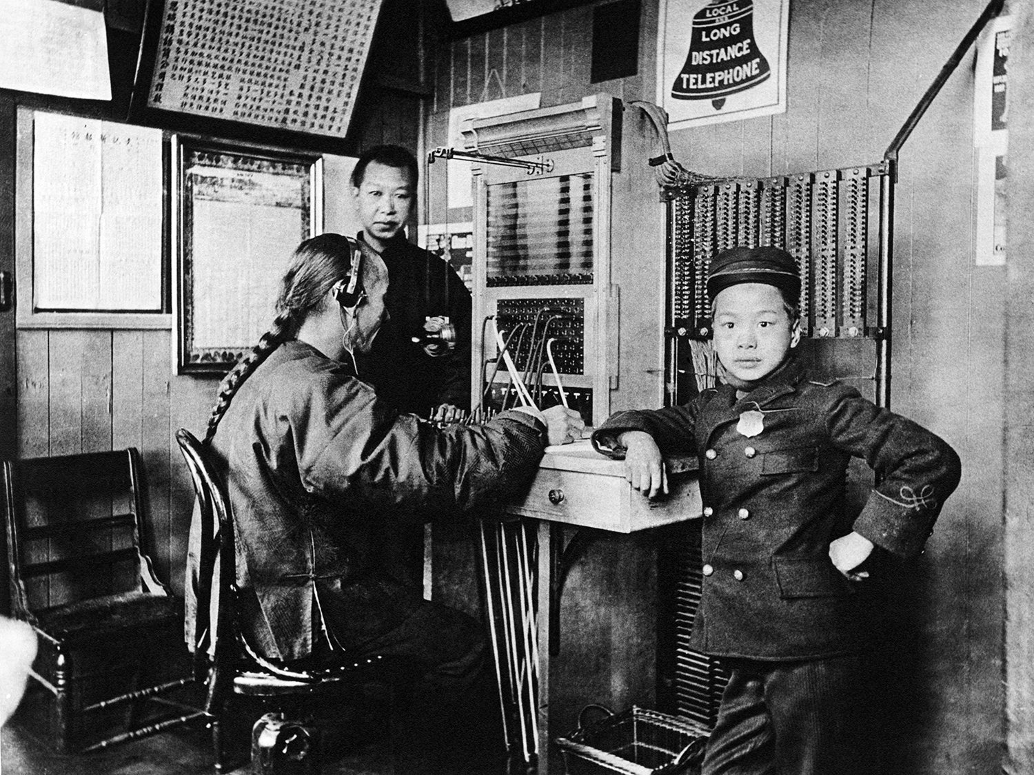 A man with a long braid wears a headset as he works. A young boy leans on a sill next to him. Another man is in the background.