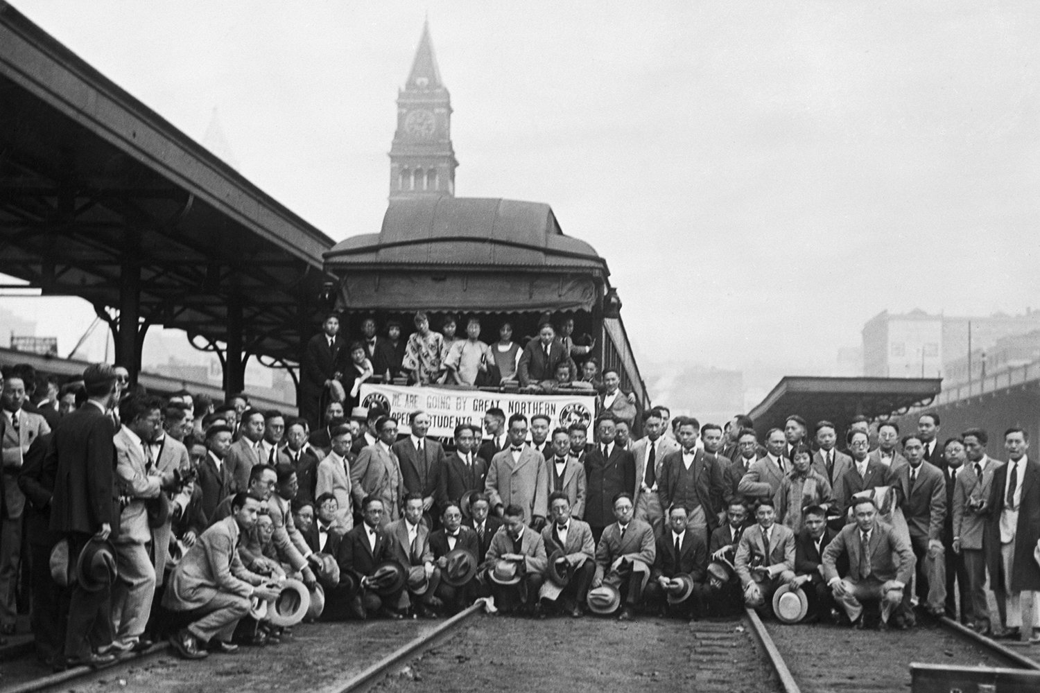 A large group of students pose for a group photo on a train platform with a clock tower in the background behind them.