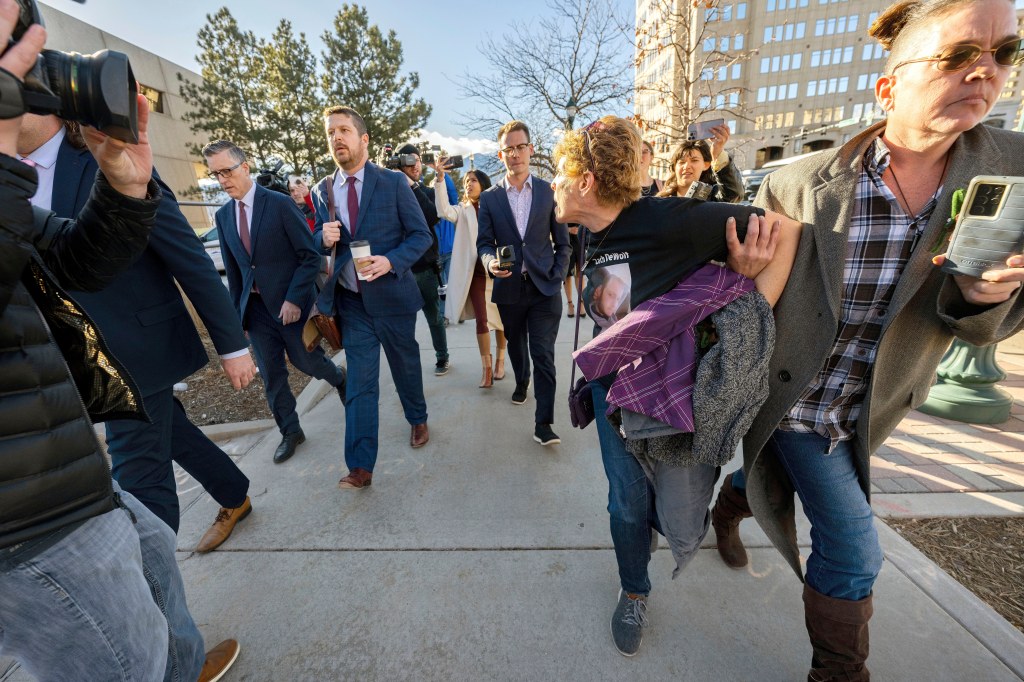 A woman being supported by another woman, walking away from a group of people in suits and media personnel.