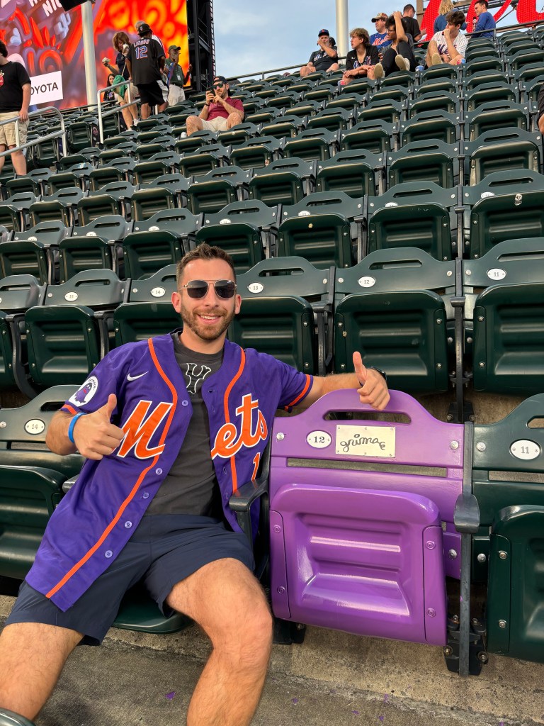 Man in Mets jersey giving thumbs up in stadium seat.