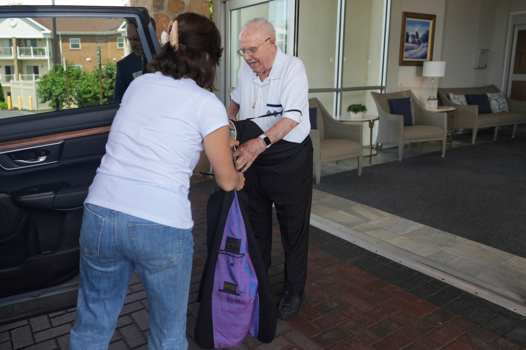 A 100-year-old priest having his jacket put on by a woman before getting into a car.