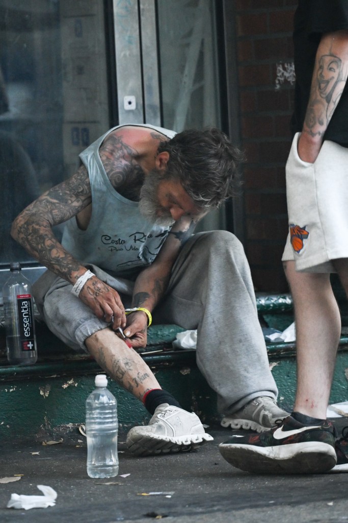 A heavily tattooed man injects his leg while sitting on the ground.