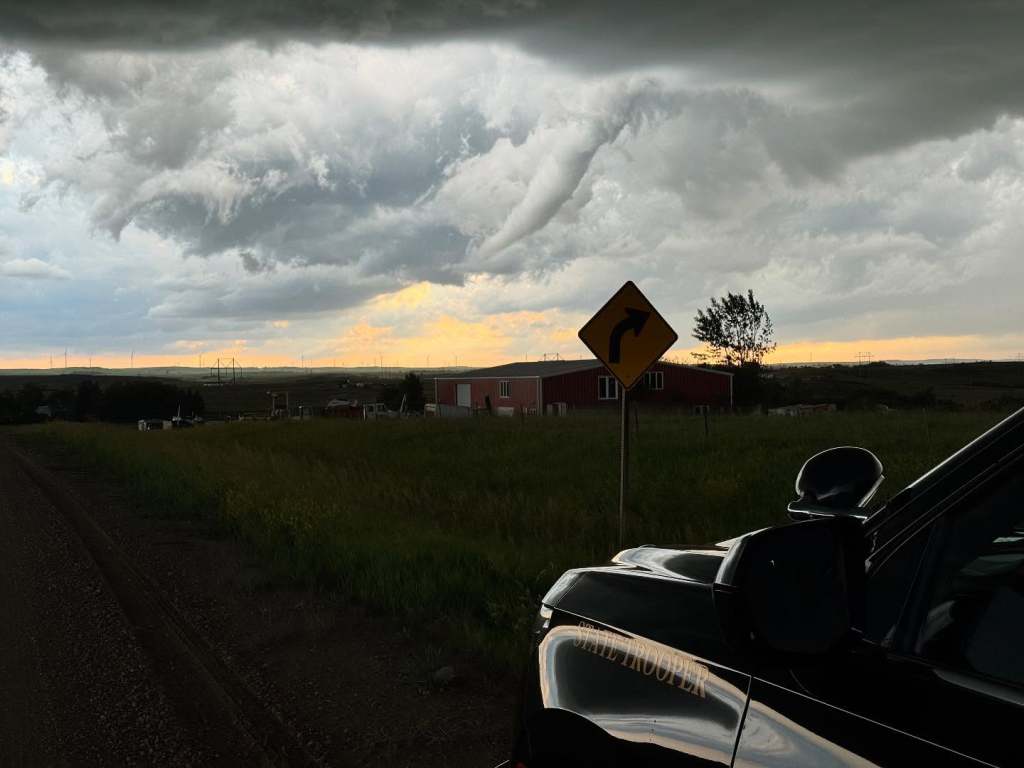 State trooper vehicle parked near a curve sign with a tornado in the background.