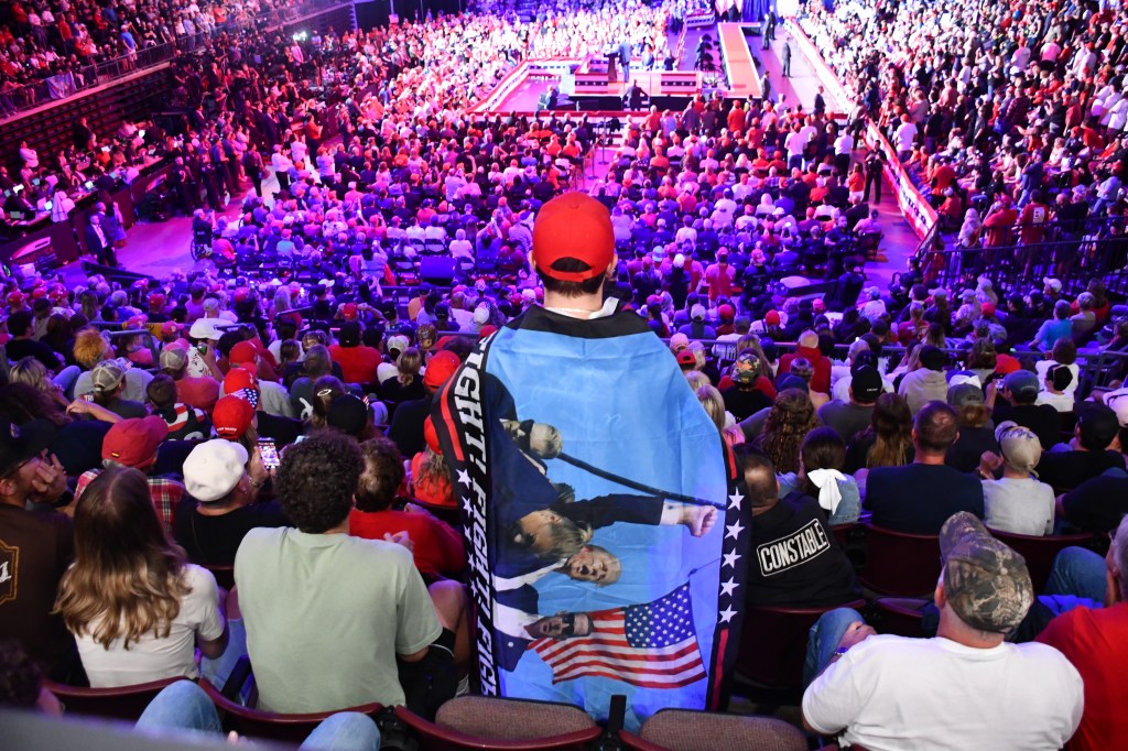 A person with a Donald Trump campaign flag draped over their shoulders sits among a large crowd of Trump supporters.