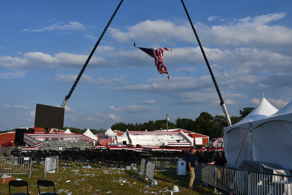 Post-event cleanup at a rally; an American flag hangs from a crane, empty chairs and litter are scattered on the ground.