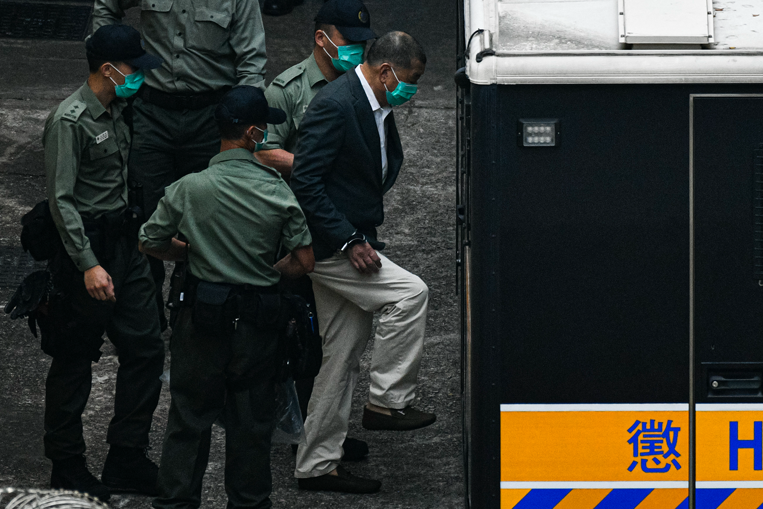Four security guards wearing matching green uniform shirts and medical face masks surround Jimmy Lai as he steps up to enter the back of a vehicle. Lai is a man in his 70s wearing khaki pants, a dark suit jacket, a face mask, and handcuffs.