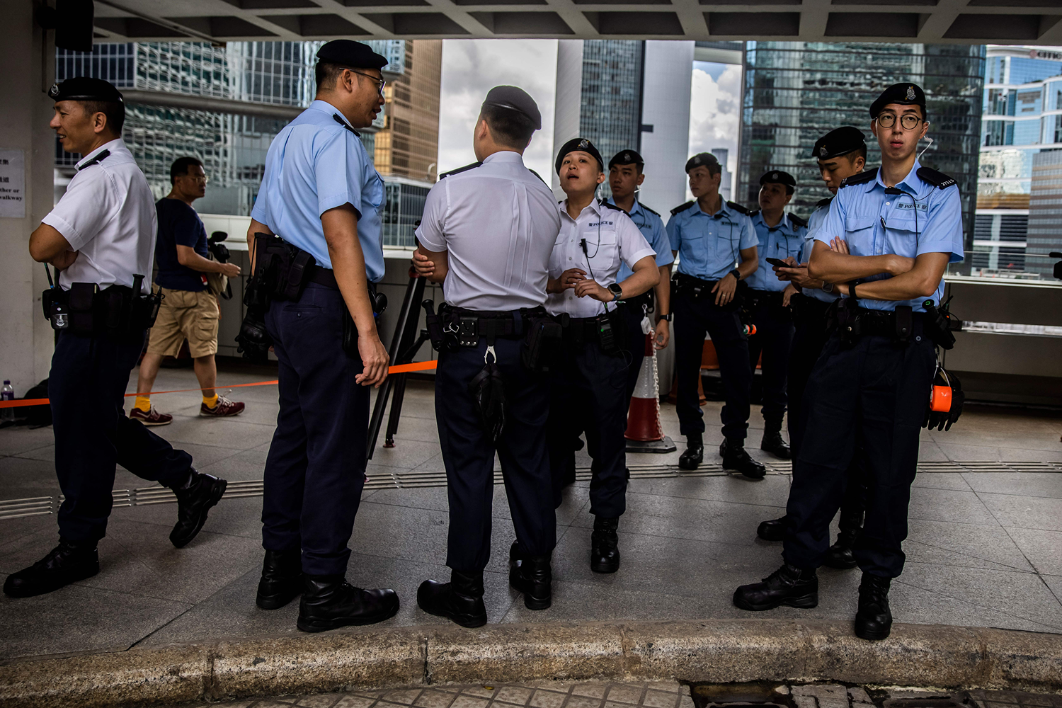 A group of police officers, wearing short-sleeved uniform shirts and matching hats, chat with each other as they stand on a sidewalk outside Hong Kong's High Court. Skyscrapers and a cloudy sky are visible behind the officers.