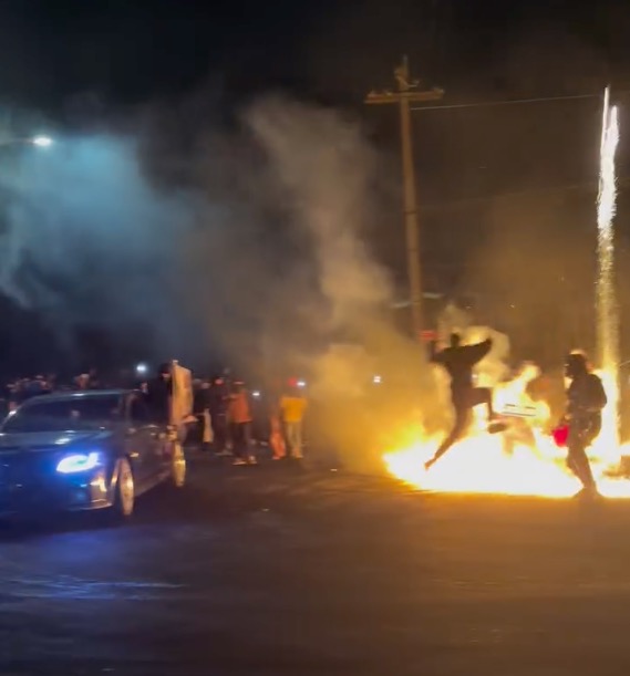 A burning car and a person jumping over flames at night.