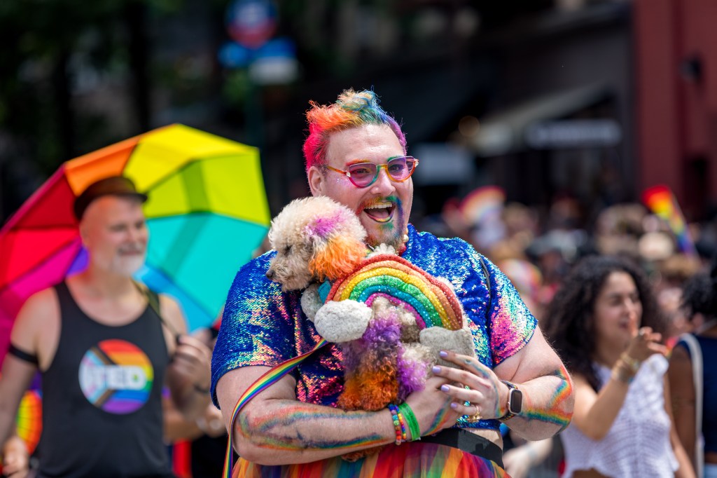 Person with rainbow-colored hair and makeup holding a dog at a Pride parade.