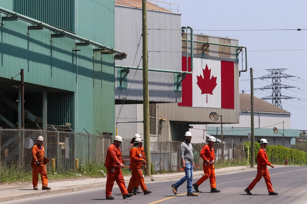 Workers crossing a street in front of a steel manufacturing plant with a Canadian flag displayed.