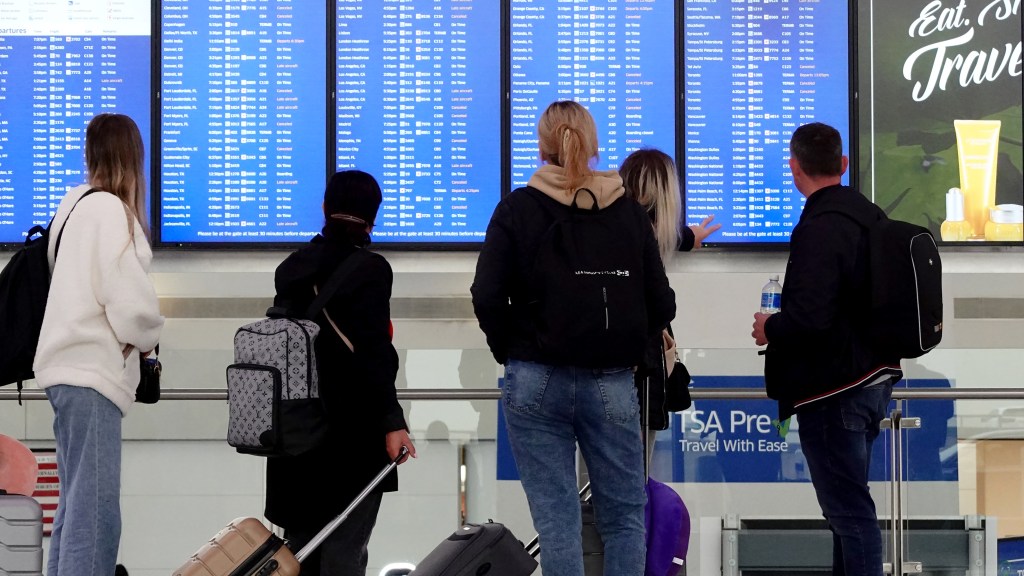 Passengers at Newark Airport check a departures board showing flight delays and cancellations.