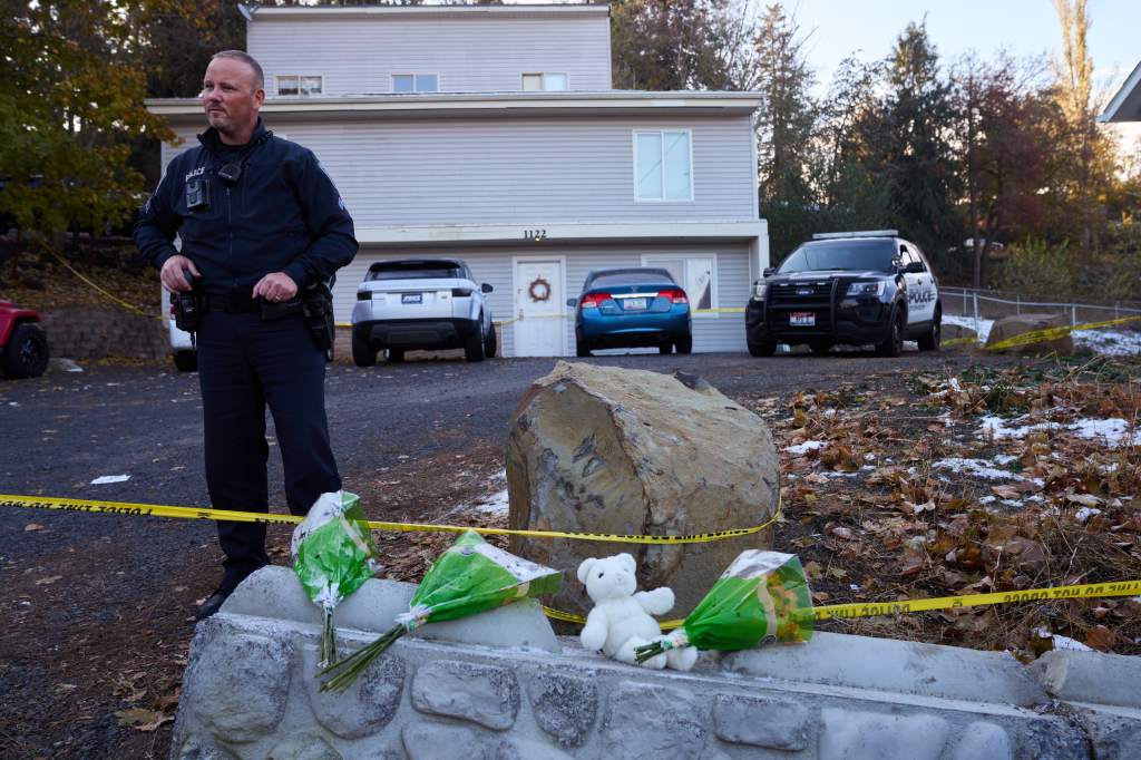 Police officer standing outside a house with crime scene tape and memorial flowers.