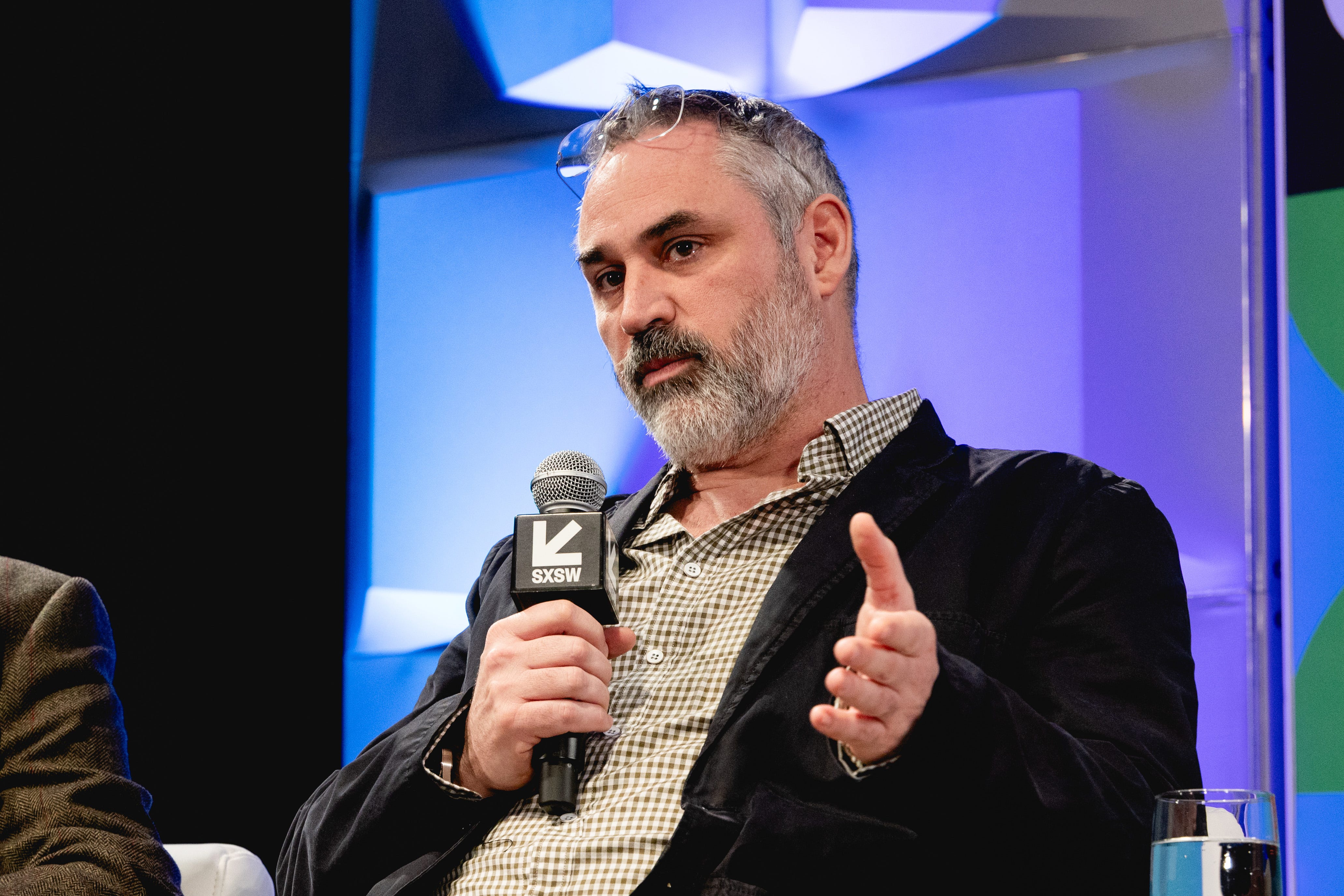 A man with short gray hair and a gray beard is holding a black microphone. Frameless glasses are resting on top of his head. He's wearing a black blazer and a checked shirt.