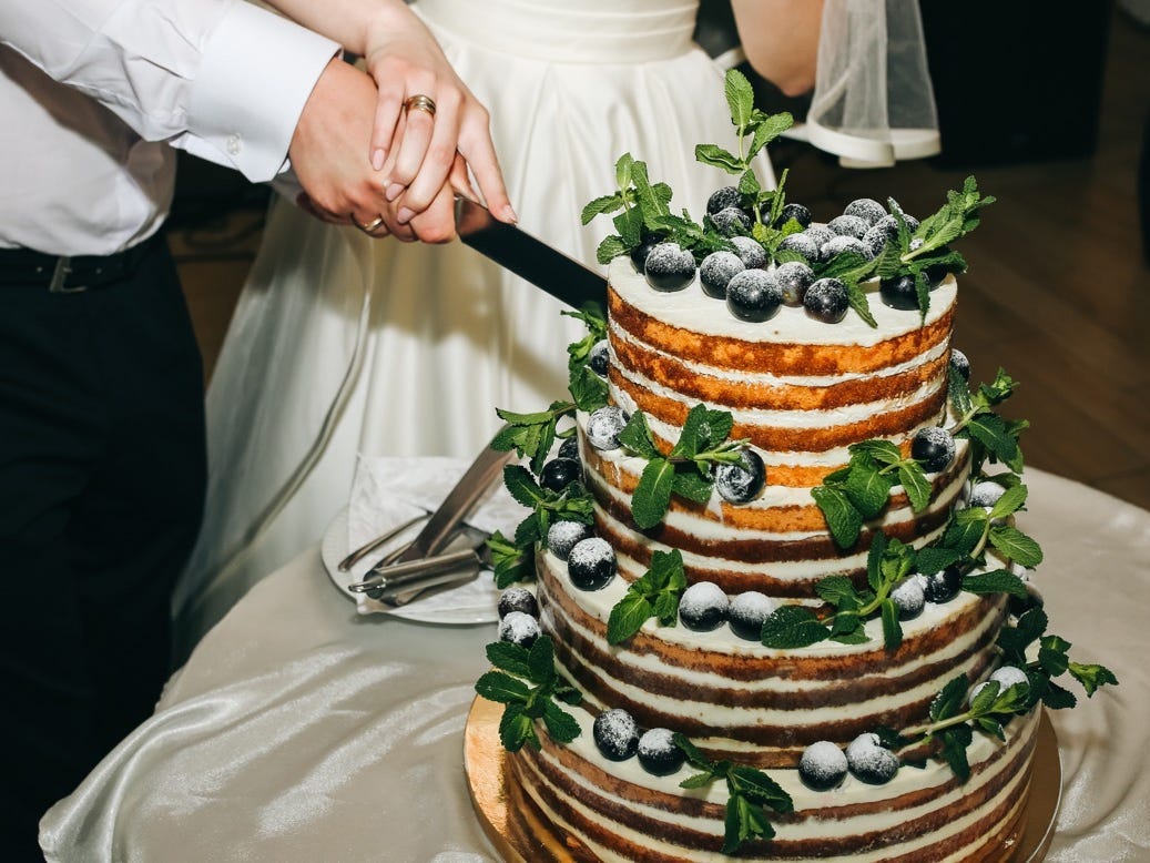 Couple holding knife together at wedding cutting a tiered cake with fruit and greenery on it