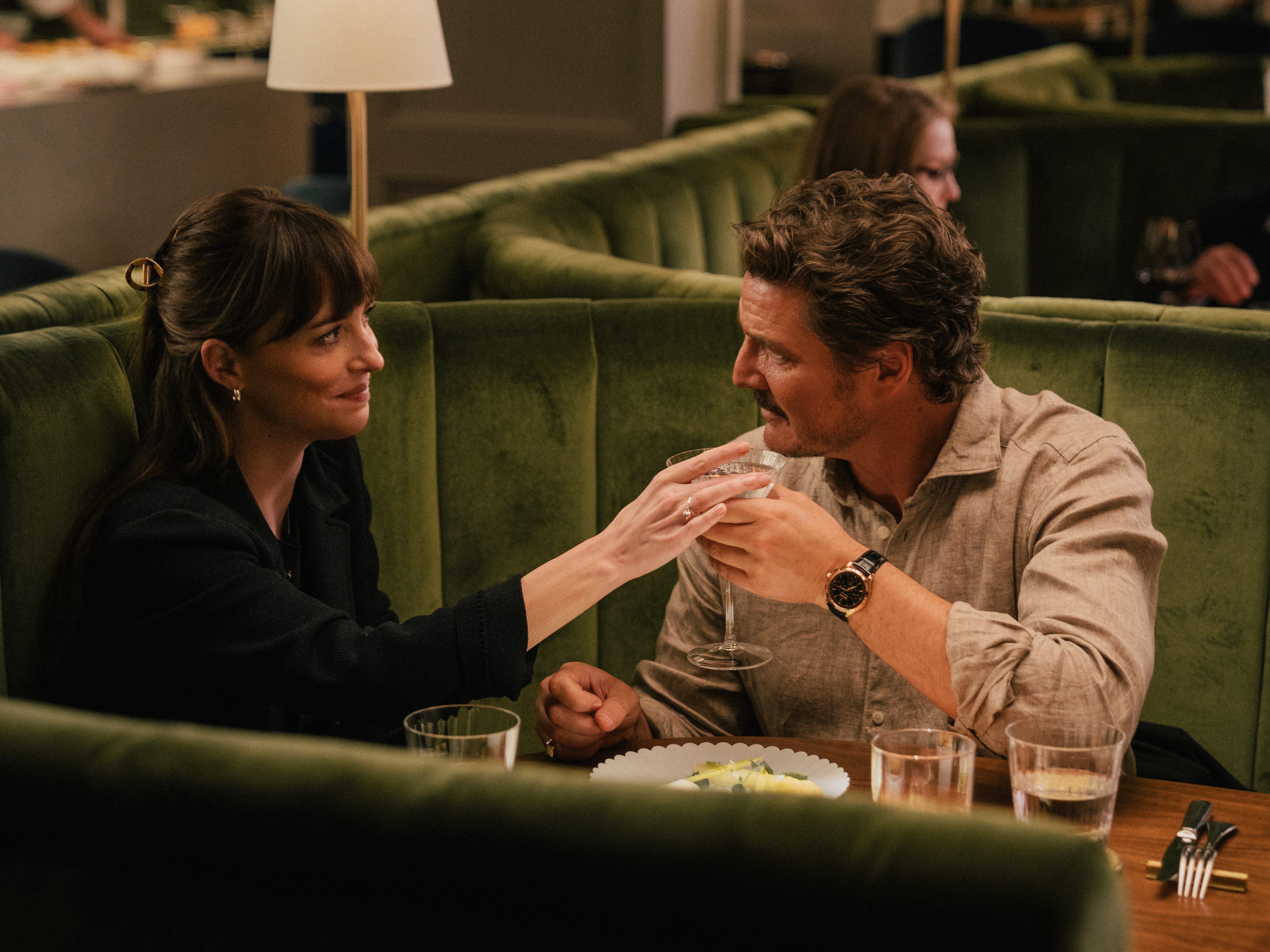 A still of "Materialists" showing Dakota Johnson and Pedro Pascal sitting in a booth and passing a cocktail glass to each other.
