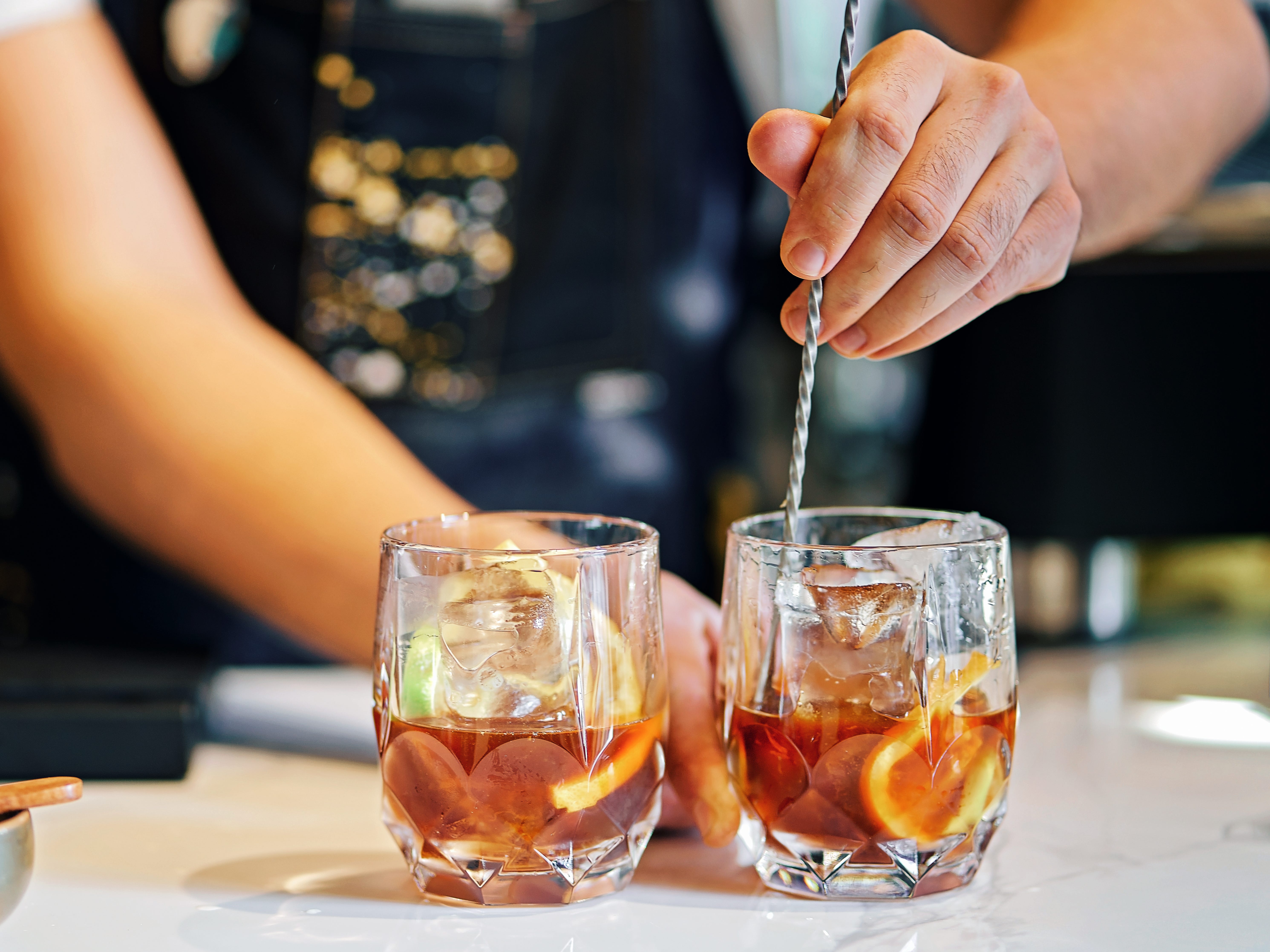 A bartender mixes two colorful cocktails with a spoon.