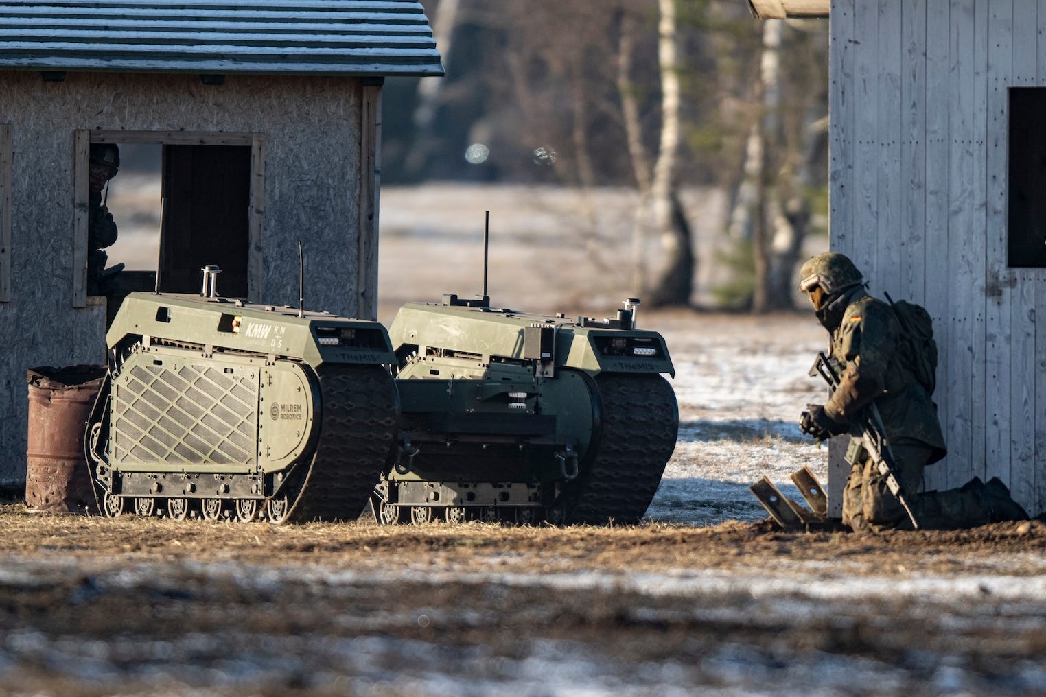 A green robotic vehicle with tracks beside two small wooden buildings and a kneeing individual