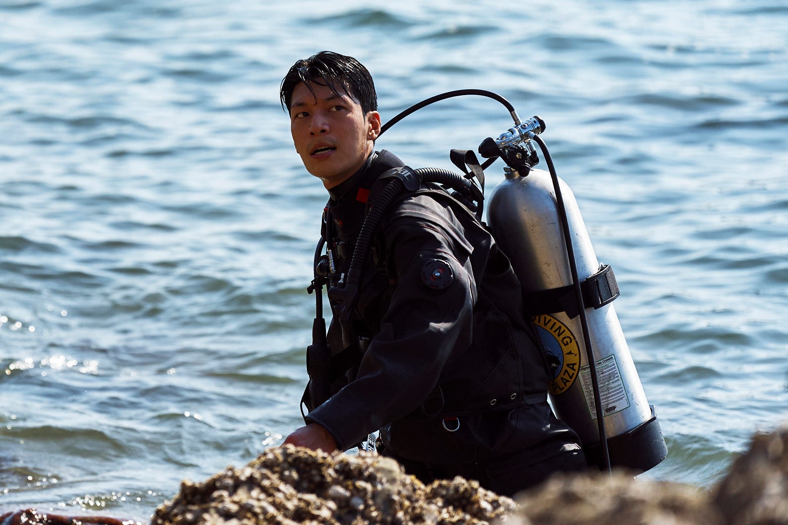 A still of "Squid Game" showing Wi Ha-jun in diving equipment walking out of the sea into rocky terrain.