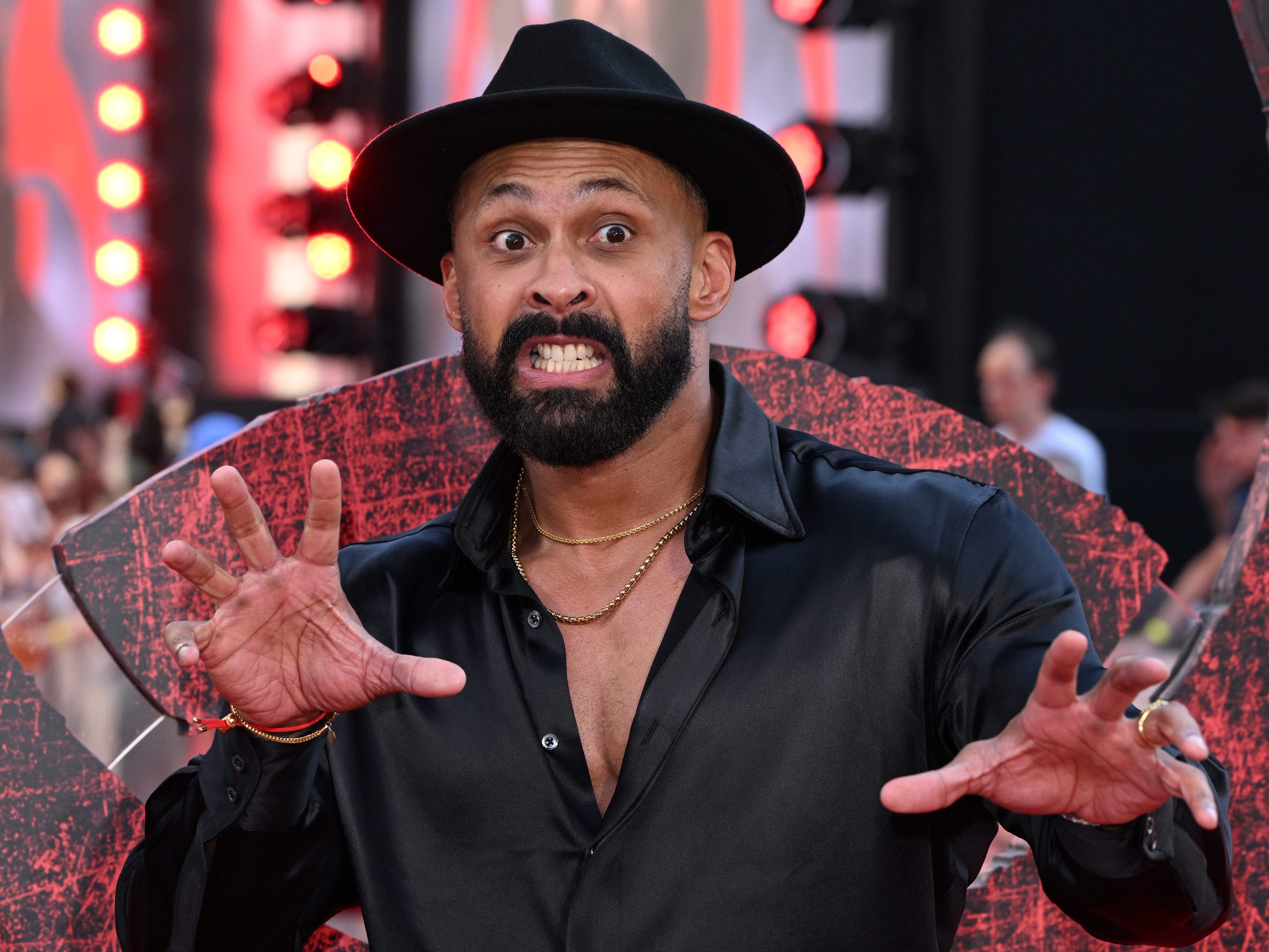 A brown man with a black beard wearing a silk black shirt with an open collar and gold chains around his neck. He's wearing a black trilby and his pretending to pull a scary face.