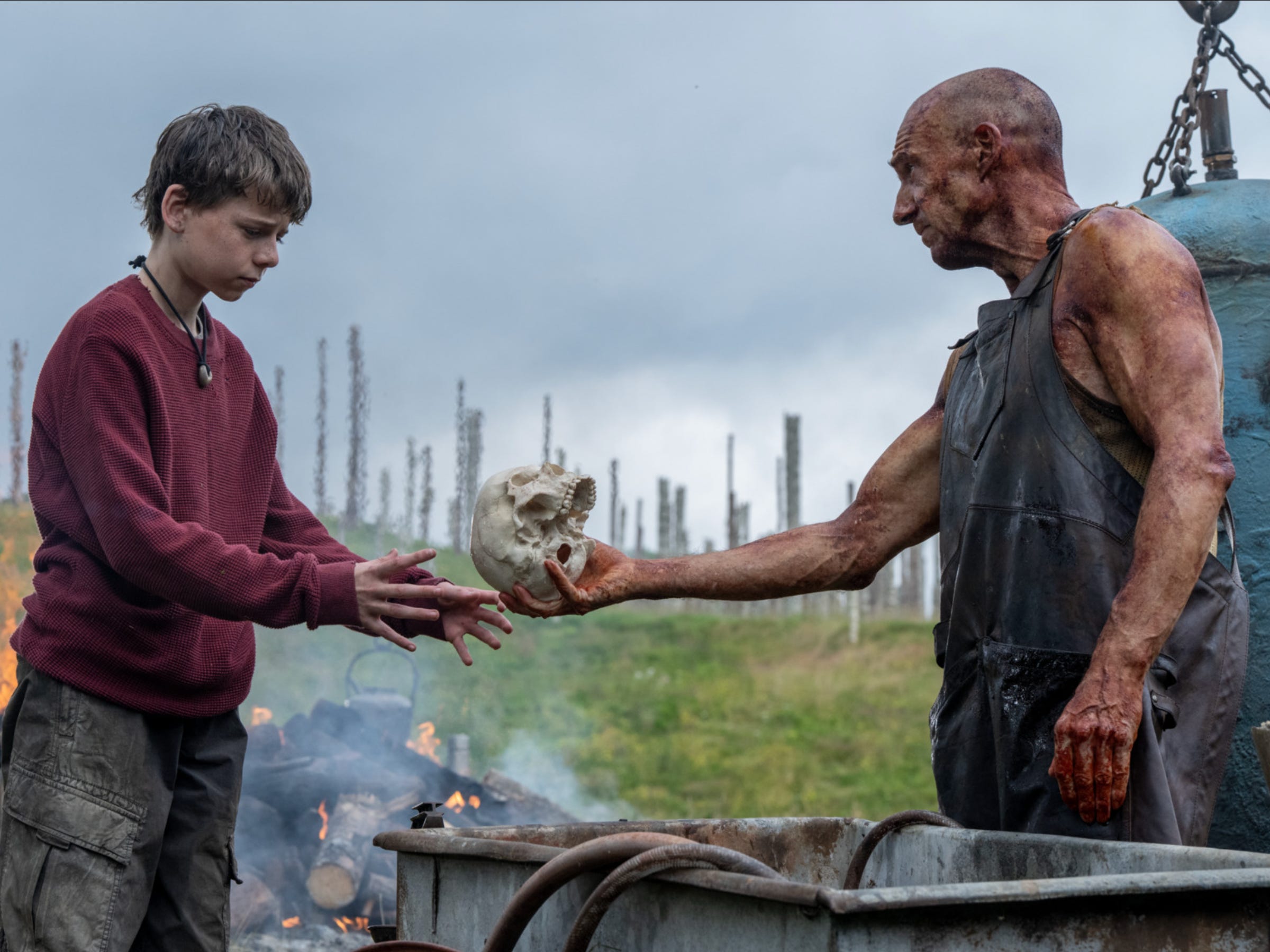 An older bald man hands a teenage boy a skull. On the left, the teenager is wearing a red sweater with a string necklace and plaid cargo pants. On the right, the older man is wearing a black apron and is handing the skull over to the boy with his right hand. Behind them, there is a pile of burning logs.