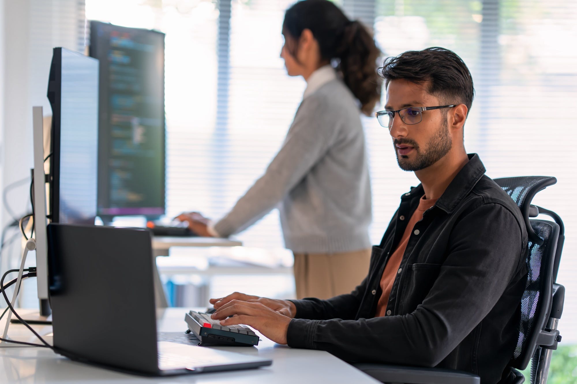 Two tech workers in an office type on keyboards in front of laptops and computer monitors.
