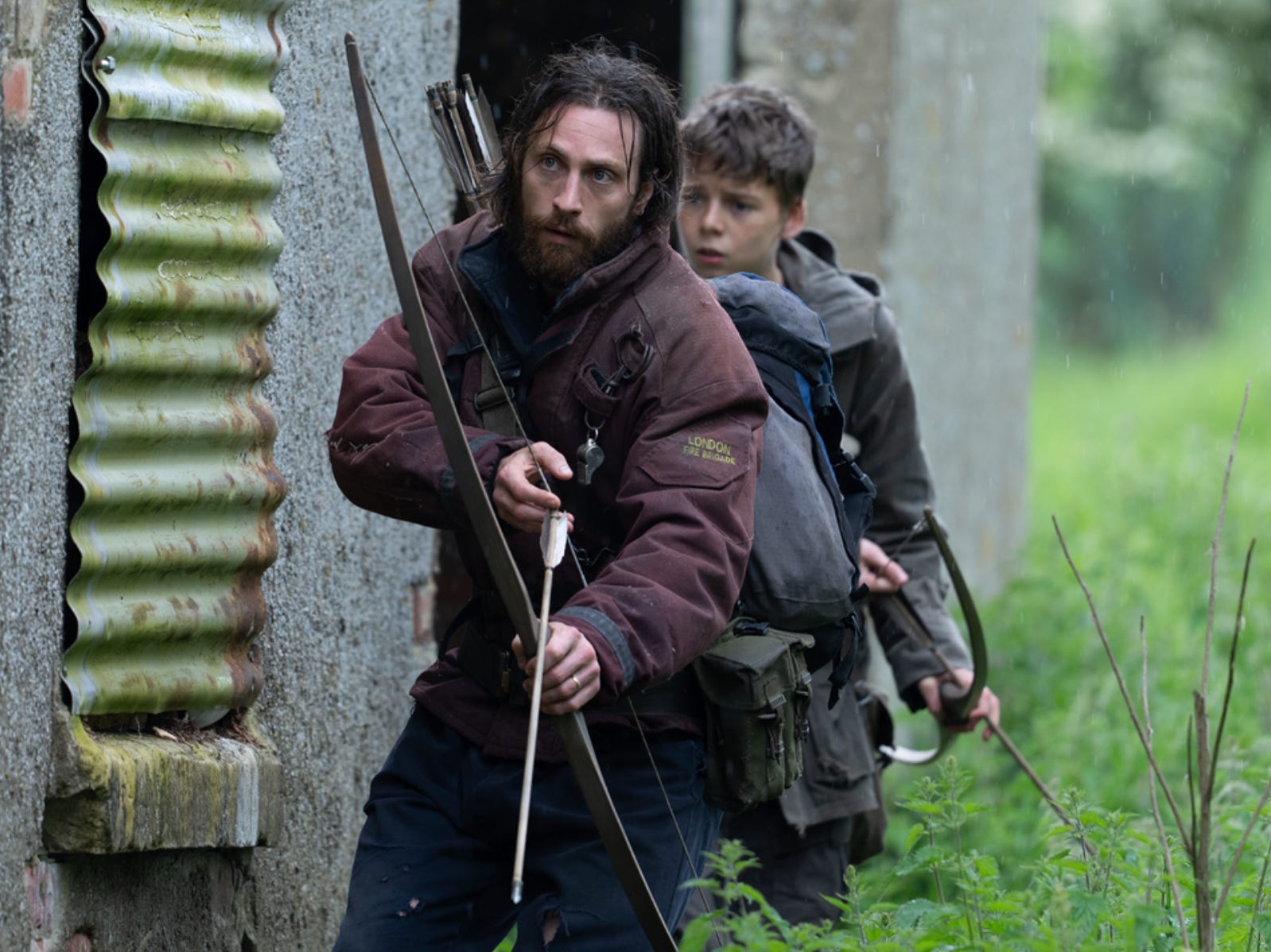 A man and his teenage son walk around the back of a concrete building that is surrounded by nettles. They're both holding bows and arrows. The man has long brown hair and a beard; he's wearing a burgundy coat with a patch that reads 