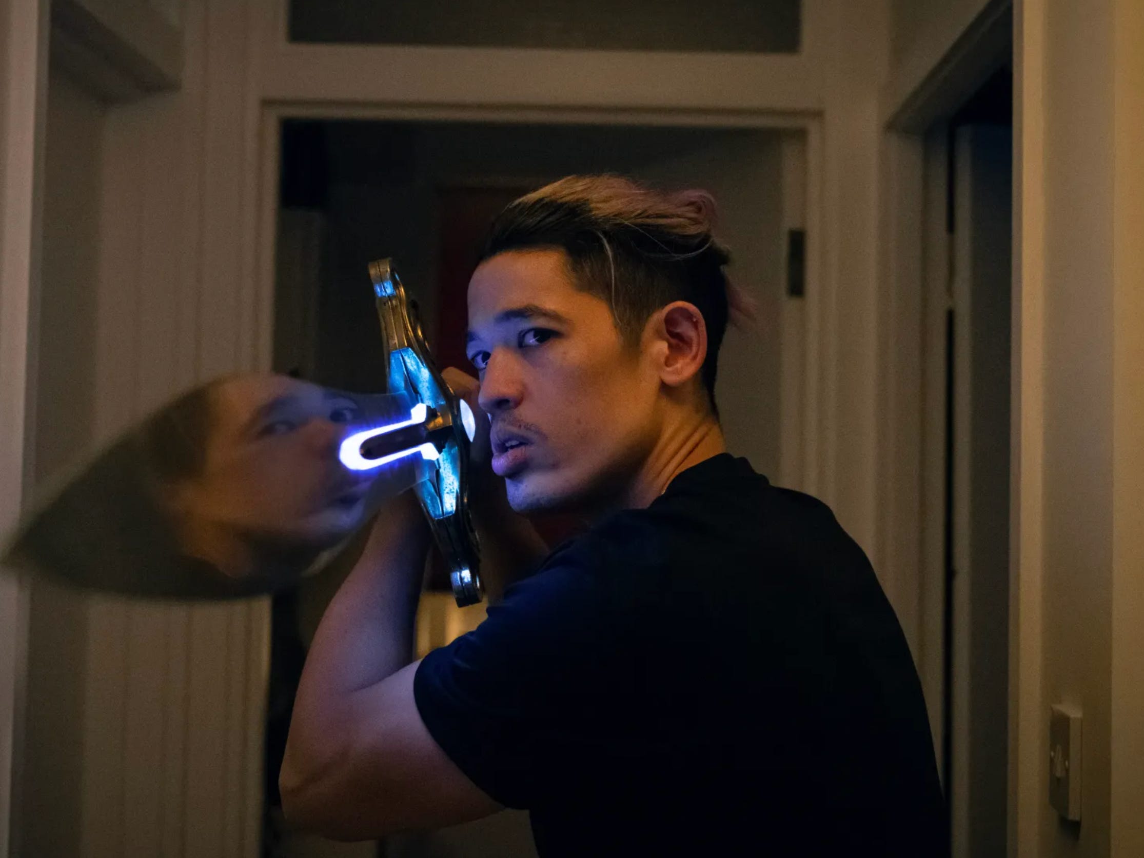 A man with slicked back black and blond hair and a short dark mustache holds a metal sword in the corridor of a flat. The sword has electronic blue lights on it.