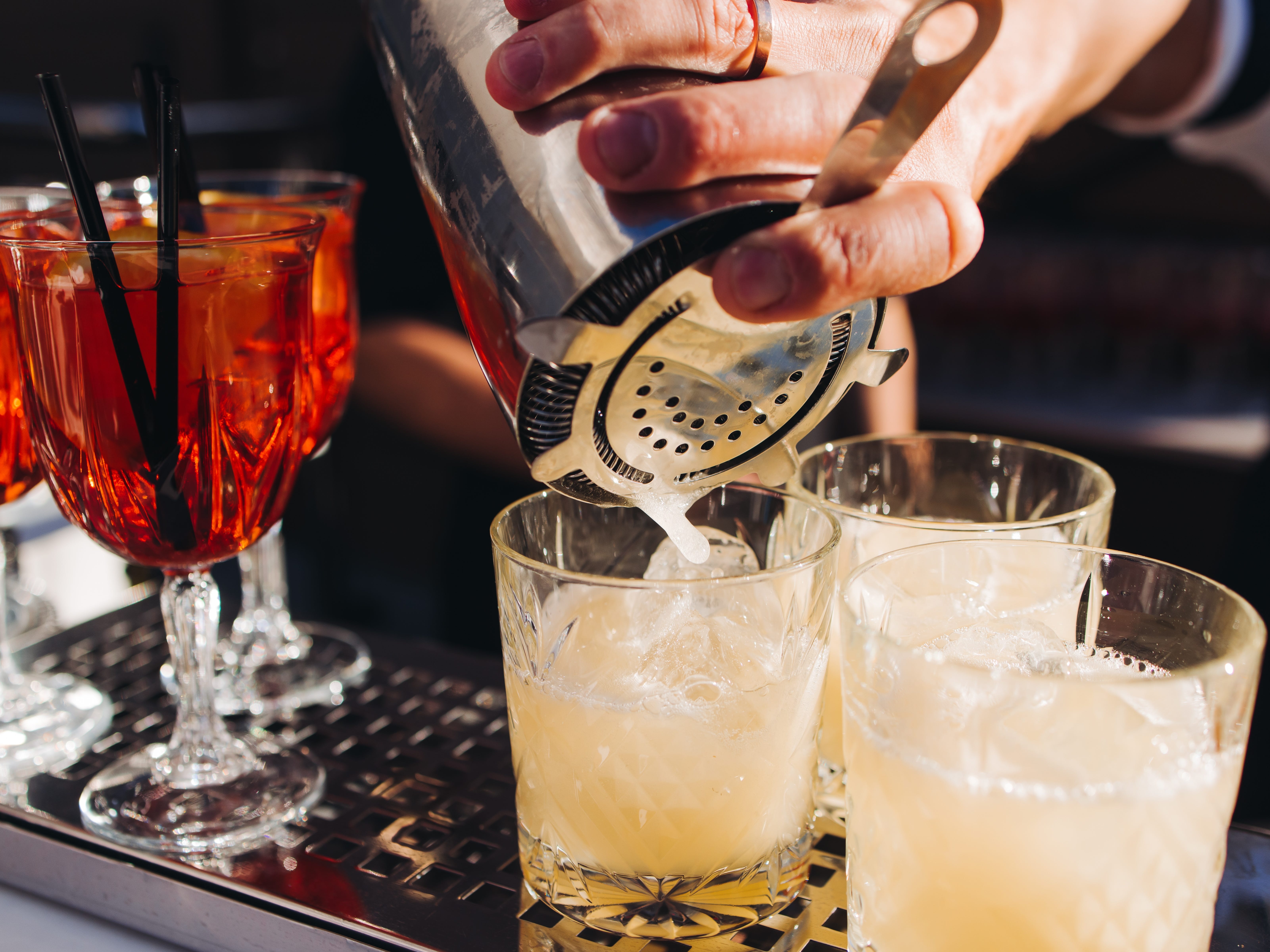 The hand of a bartender pouring a drink from a cocktail shaker into three short glasses. Glasses of another cocktail are also on the bar.