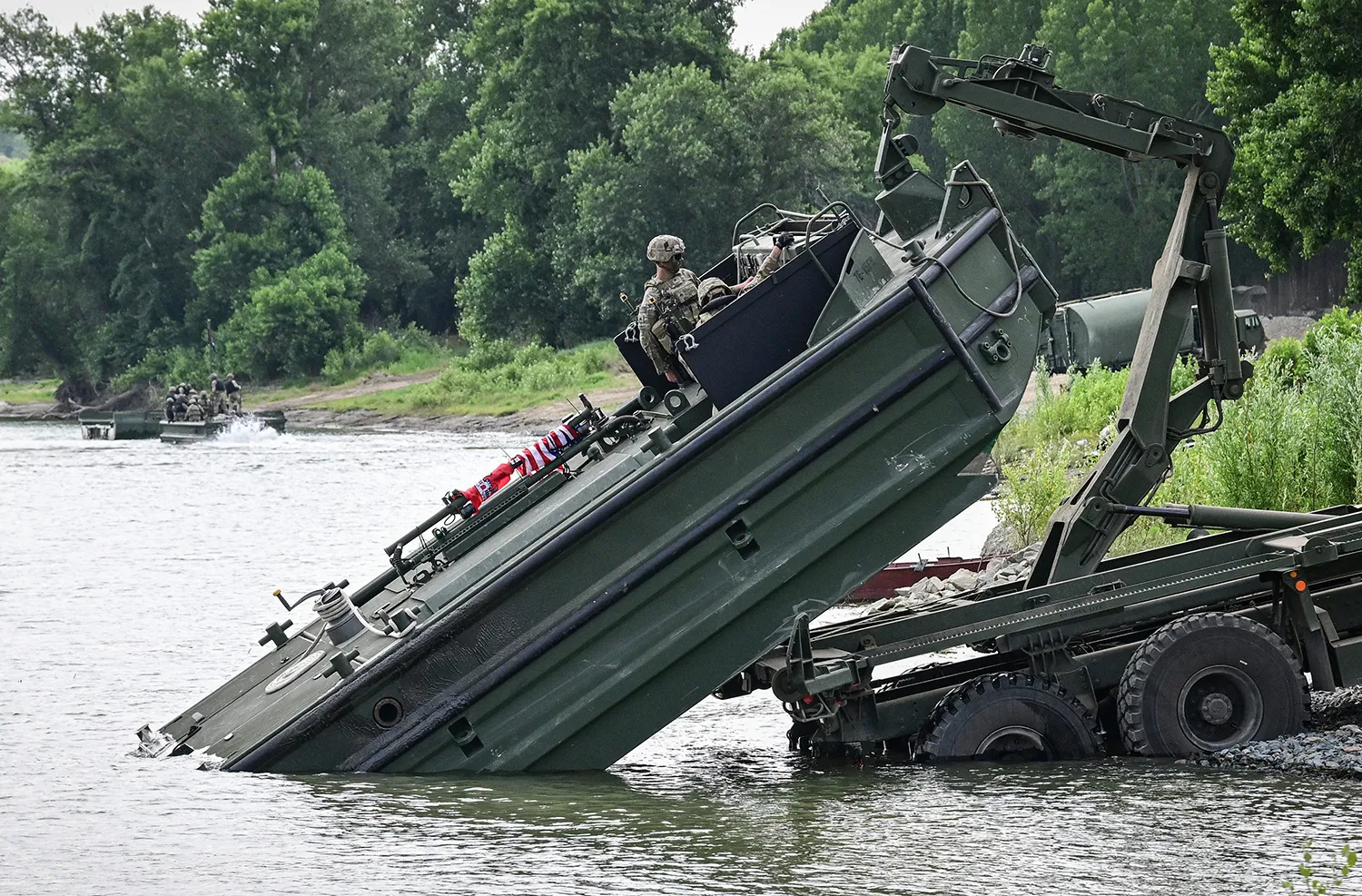 A wheeled piece of military equipment pulls a floating metal platform from the water. Soldiers are seen at the controls.