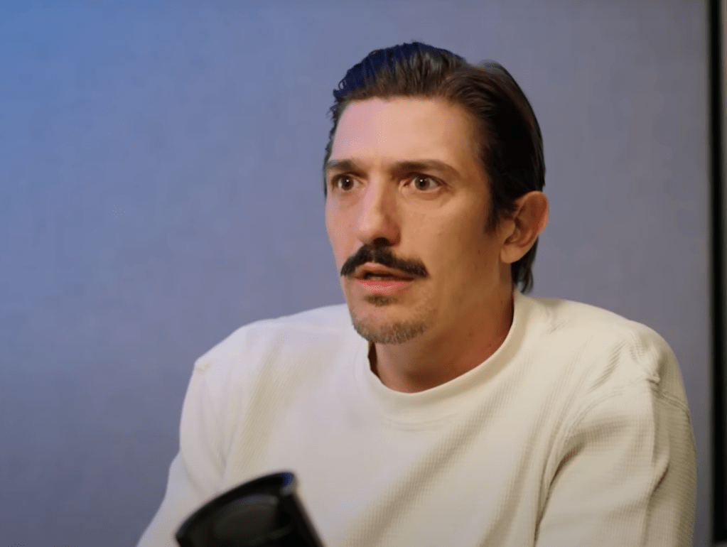Man with mustache speaking into microphone.