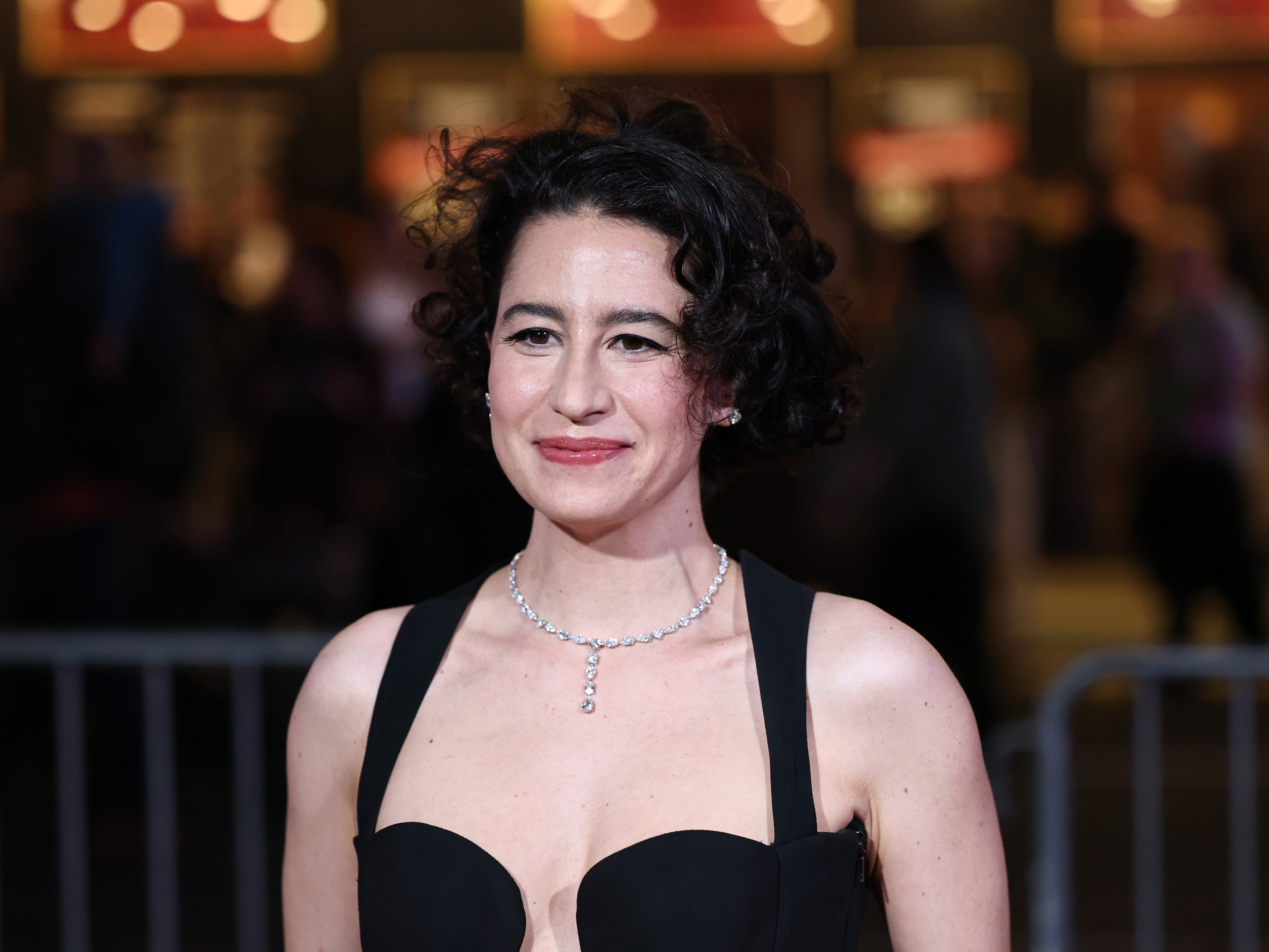 Ilana Glazer smiling for the camer awhile wearing a diamond necklace and a black dress.