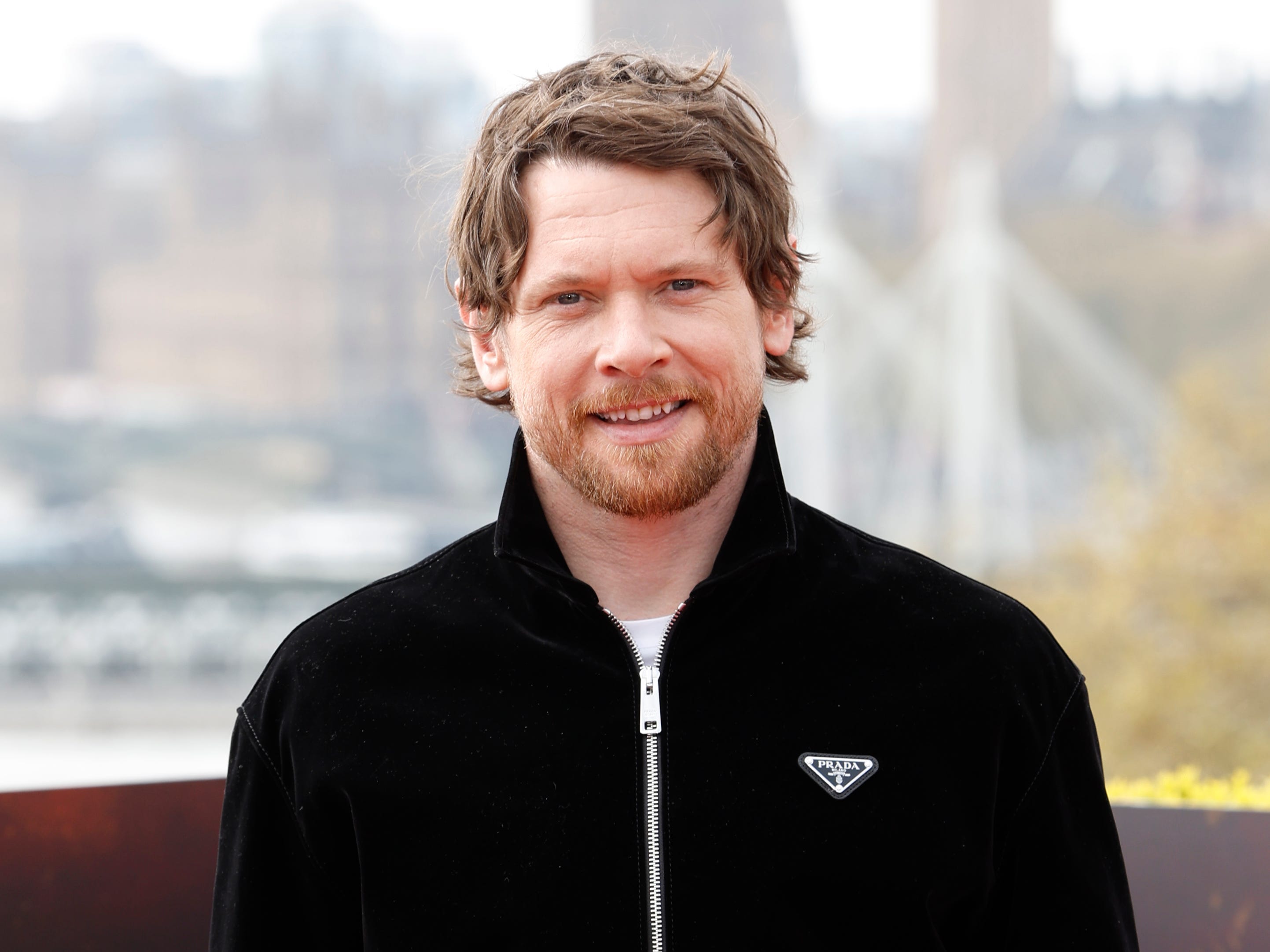 A man with brown hair and a short beard smiles at the camera. He's wearing a black sports jacket with a triangular logo that reads