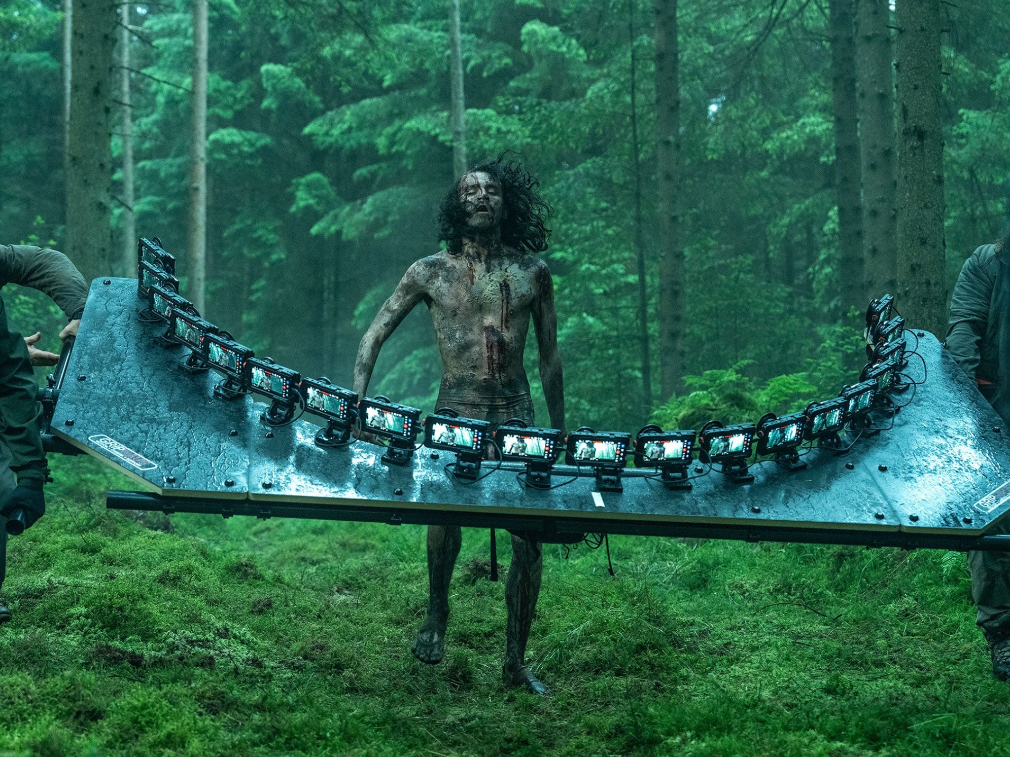 A group of men carry a curved metal plate through a green forest with 20 iPhones attached to it. In the middle of the plate is a skinny man covered in mud with long, dark hair.