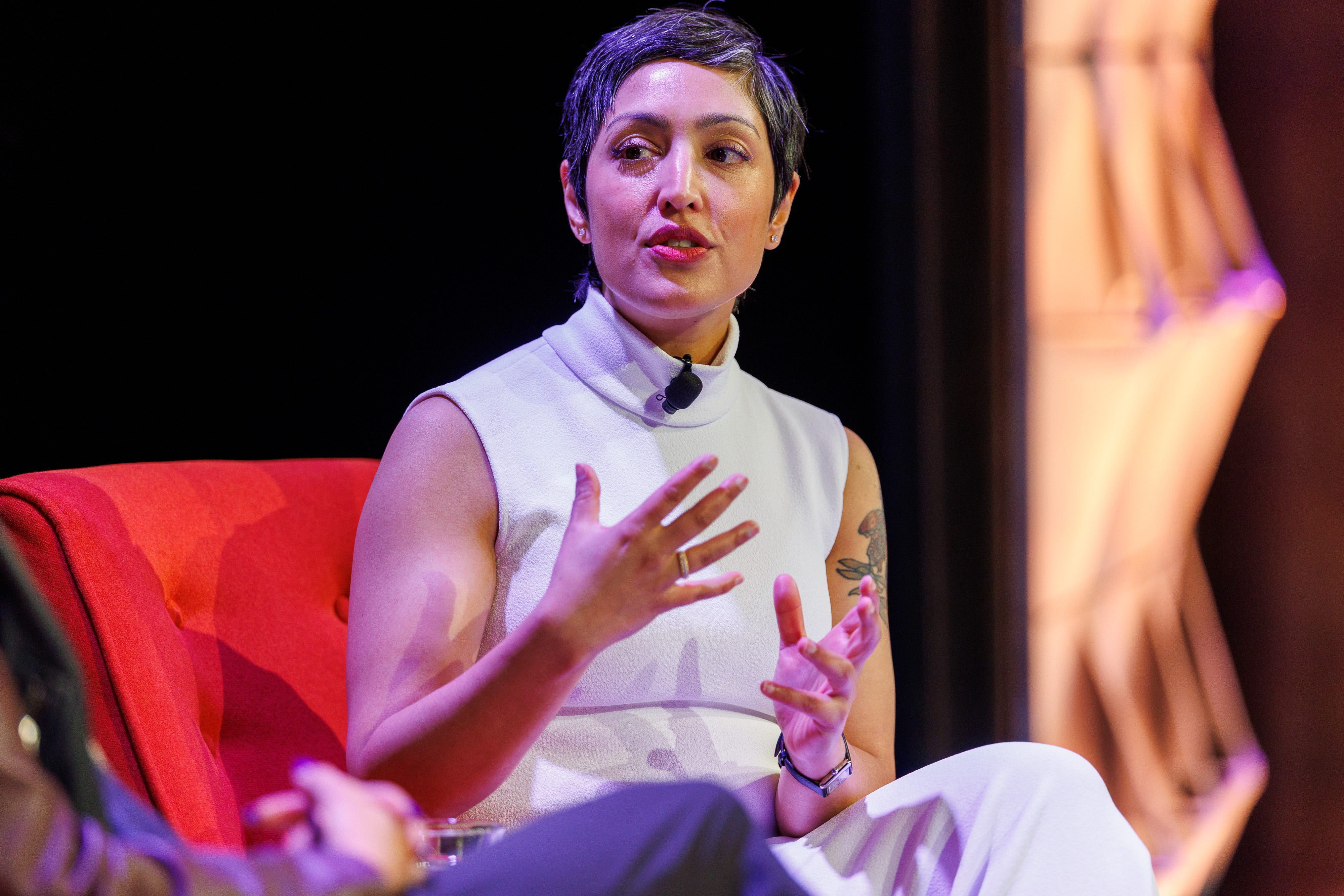 Dr. Rumman Chowdhury, CEO of Humane Intelligence, sits in a red chair, wearing a grey sleeveless turtleneck. She is mid-speech, and her arms are held slightly aloft in front of her. She is gesturing with her hands.