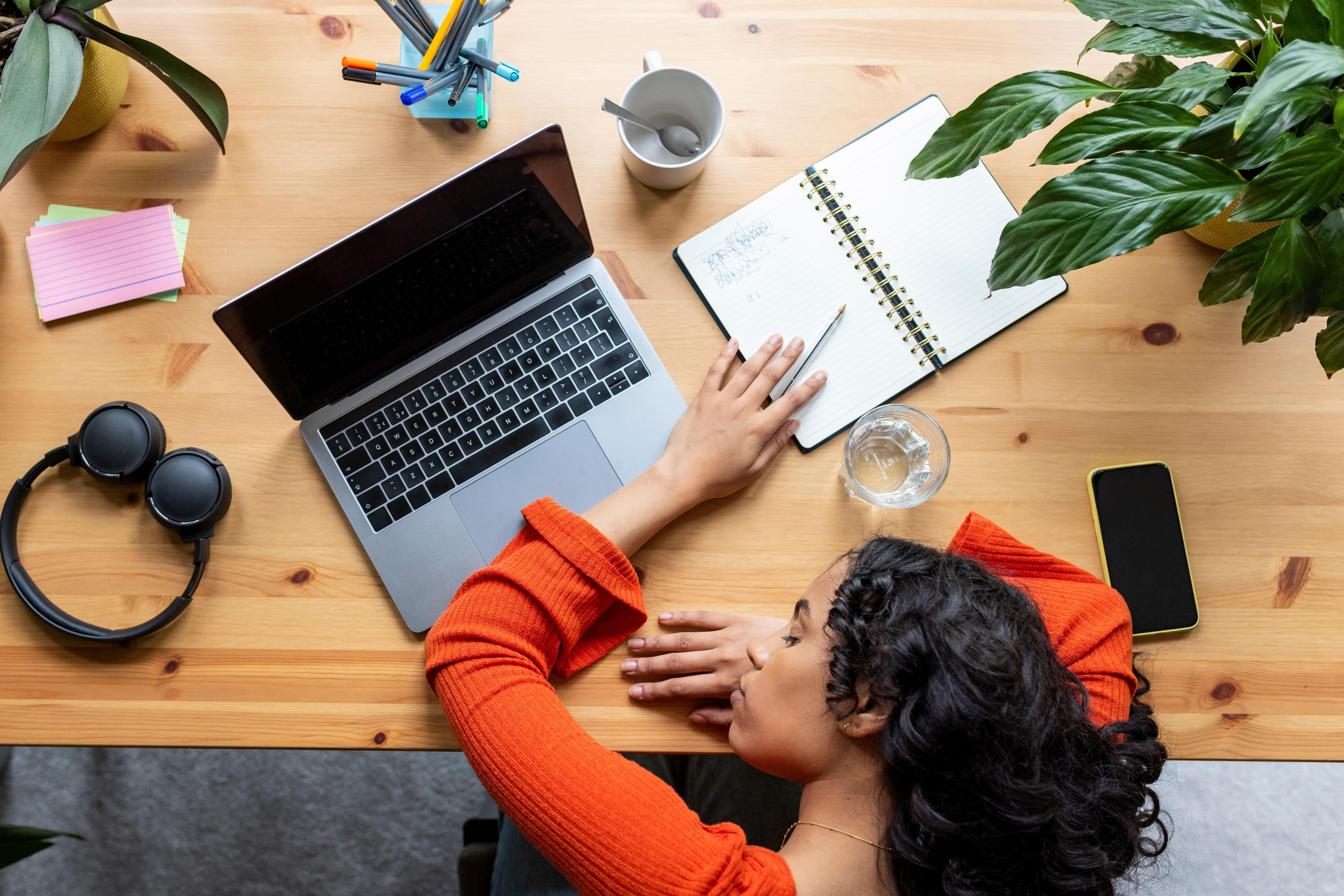 A tired woman sleeps in front of a laptop that is on top of a desk with plants.