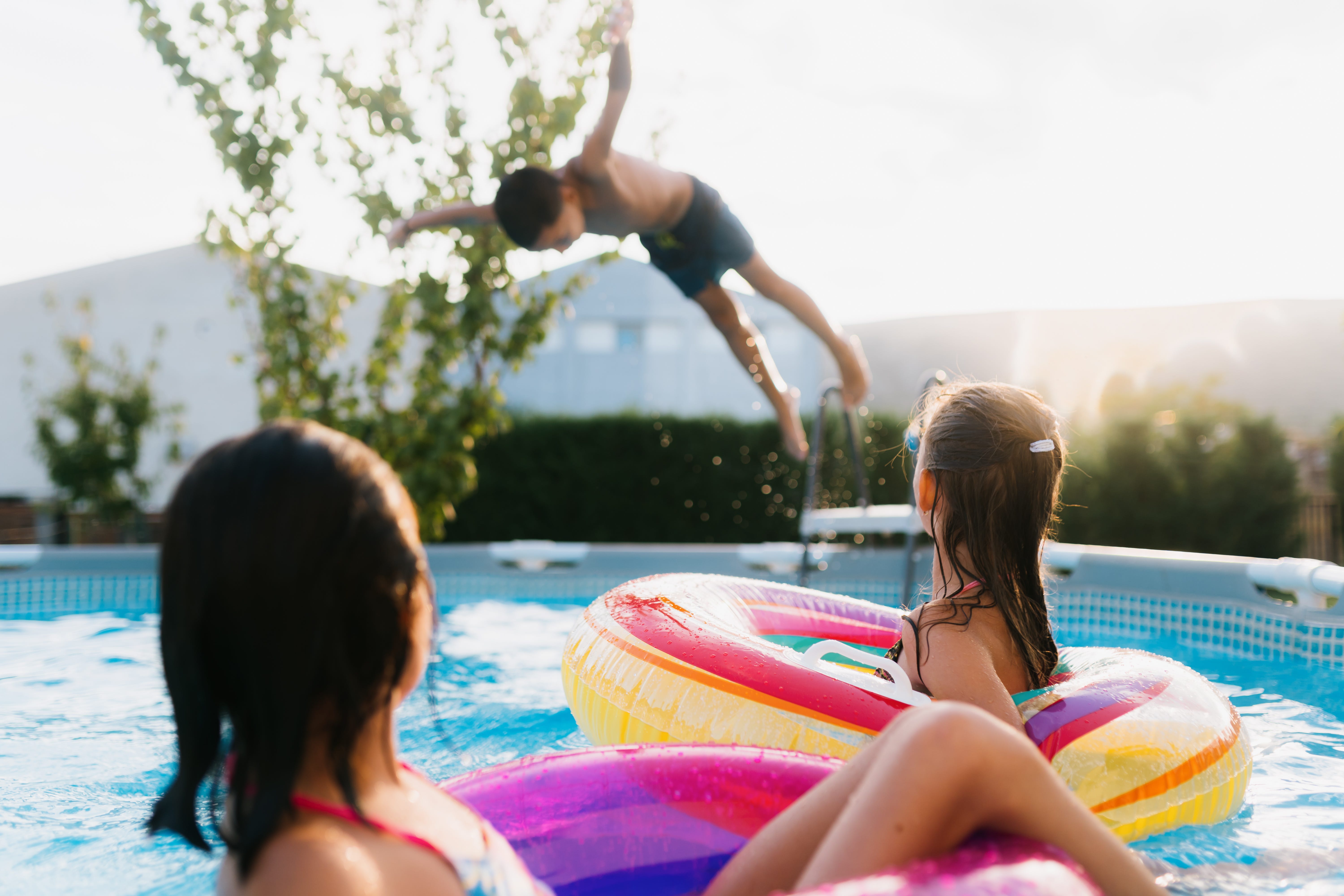 Teenage boy jumping into a pool while two young girls in flats look on.