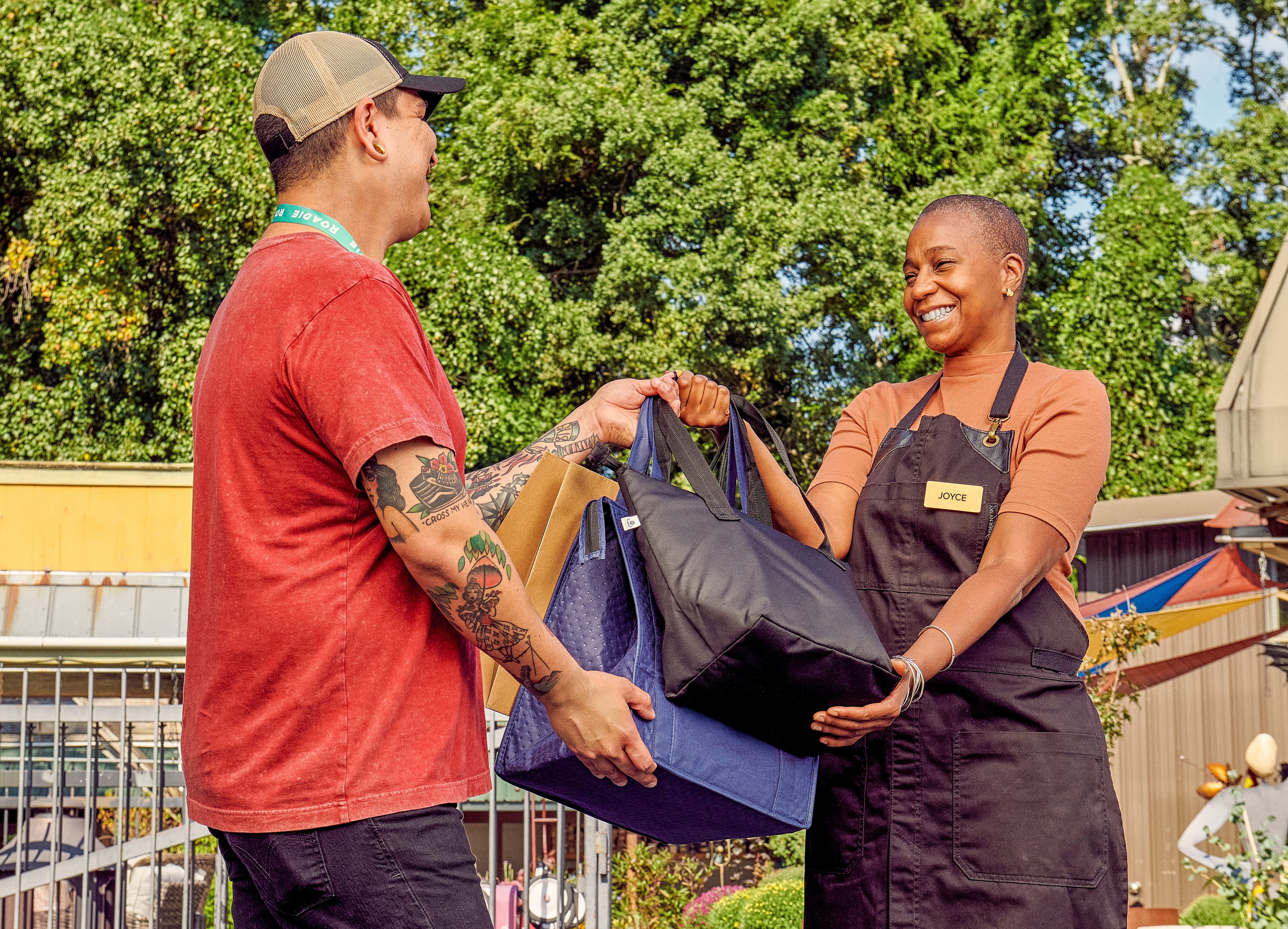 A woman wearing a nametag that reads "Joyce" hands a few bags to a Roadie delivery worker wearing a baseball cap.