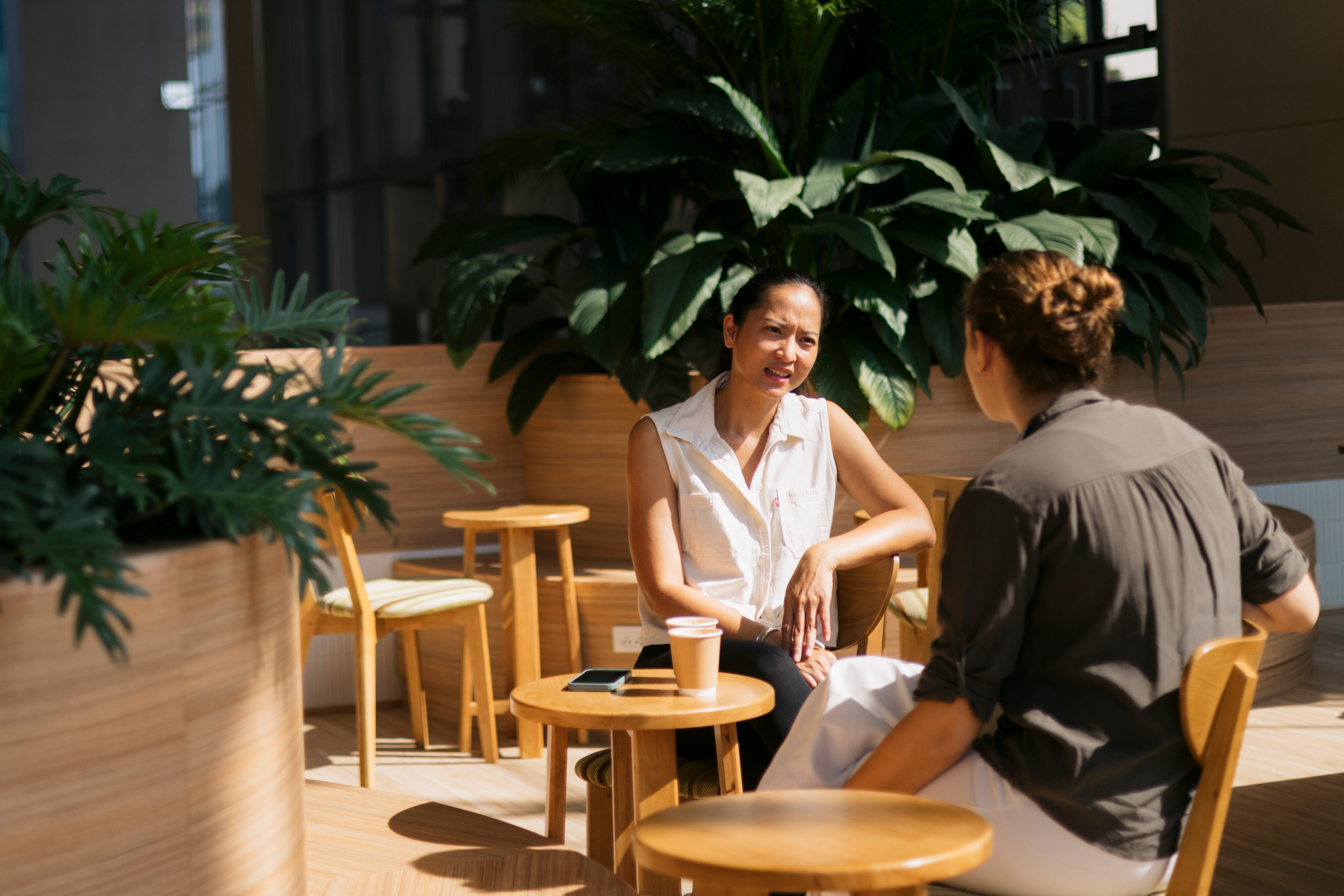 Two businesswomen enjoying a coffee break conversation in a modern office space, surrounded by lush greenery