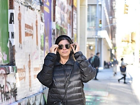 The author wearing a black puffer jacket and sunglasses and carrying a black purse, walking down a street.