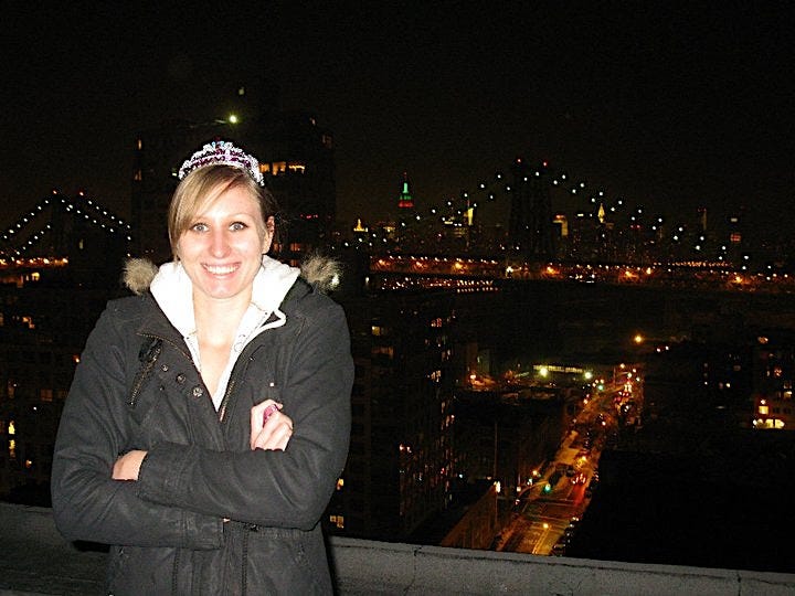 Heidi Lux standing in front of a bridge at night and smiling. There are city lights behind her and she is wearing a black jacket and a tiara and crossing her arms.