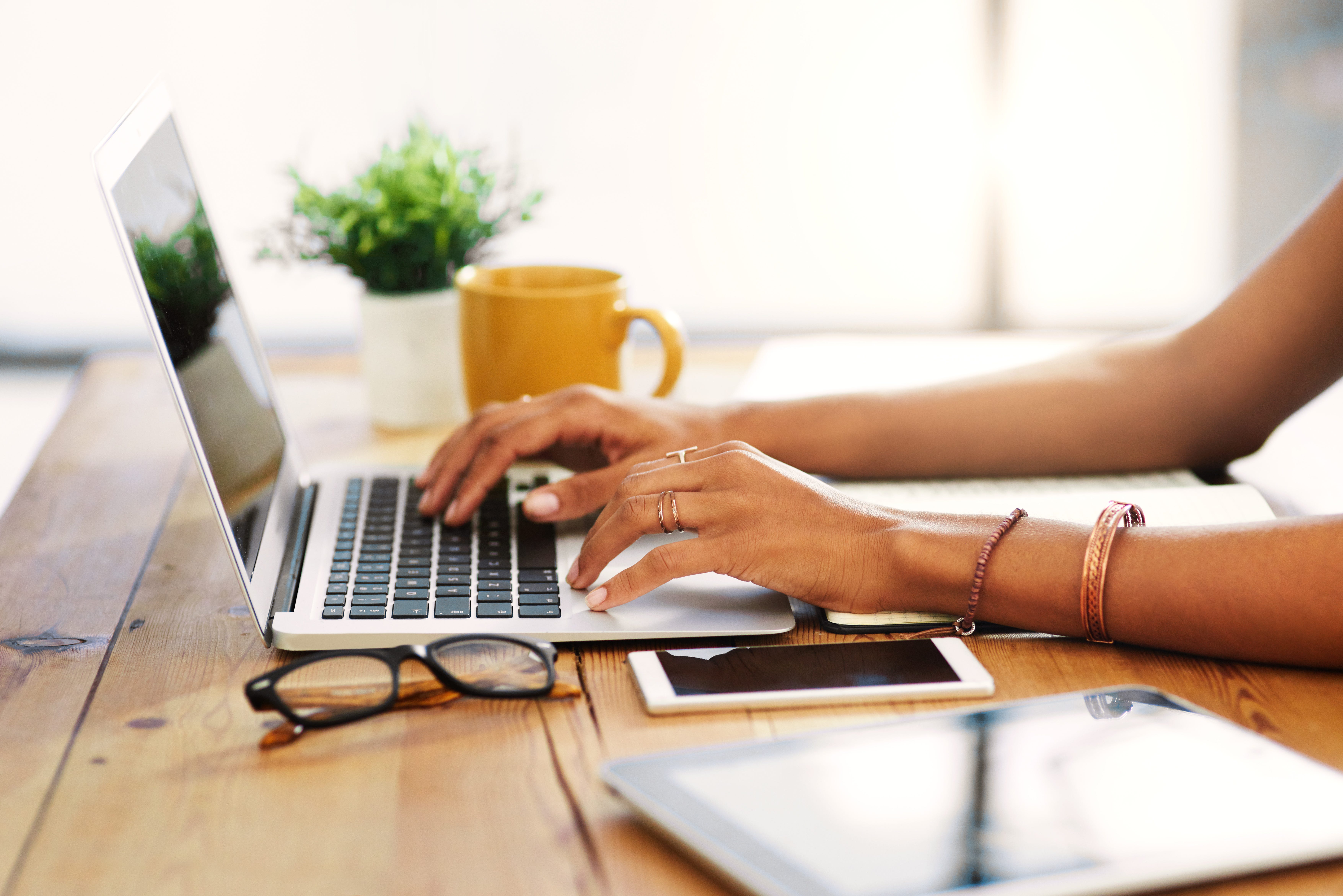 A woman working from a laptop on a wooden table, with glasses and a tablet and phone on the table. Glasses and a yellow coffee mug are also on the table.