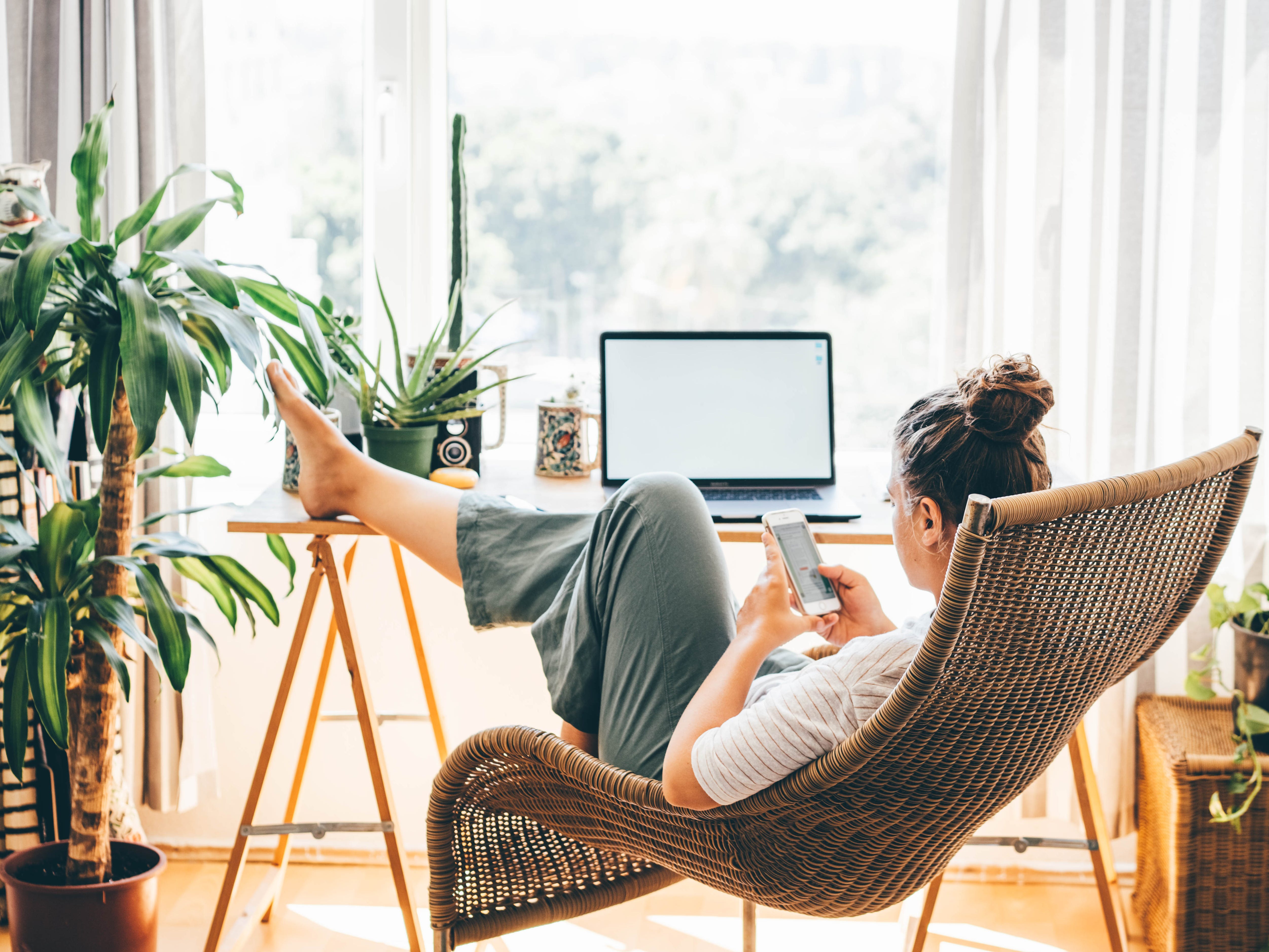 A woman reclining on a chair looking at her phone while sitting at a desk with a laptop on it.