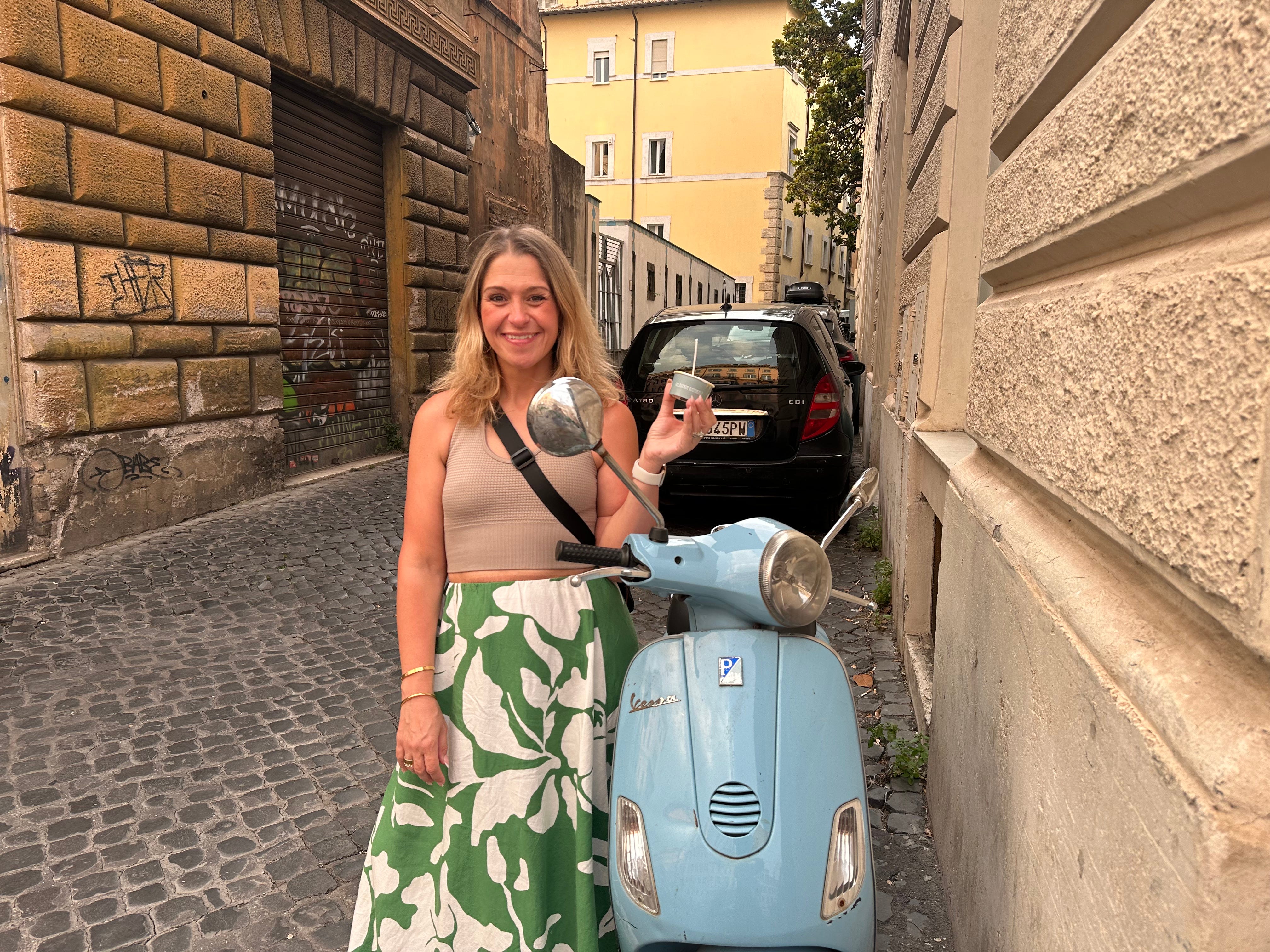 A woman standing on a cobblestoned street, next to a Vespa, holding gelato in Italy.
