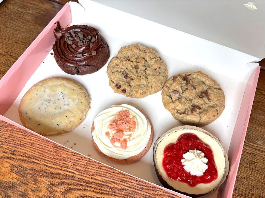 A pink box with six flavors of Crumbl cookies. The box is sitting on a wooden table.