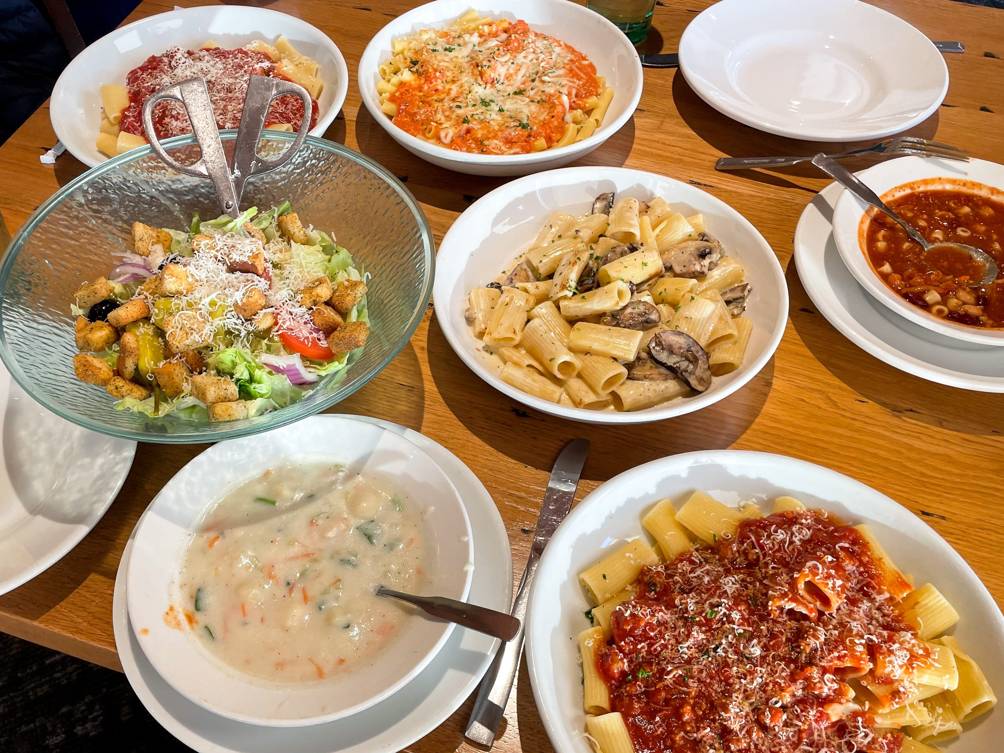 Four different pasta dishes, two different soups, a large bowl of salad, and a half-eaten breadstick on a table at Olive Garden.