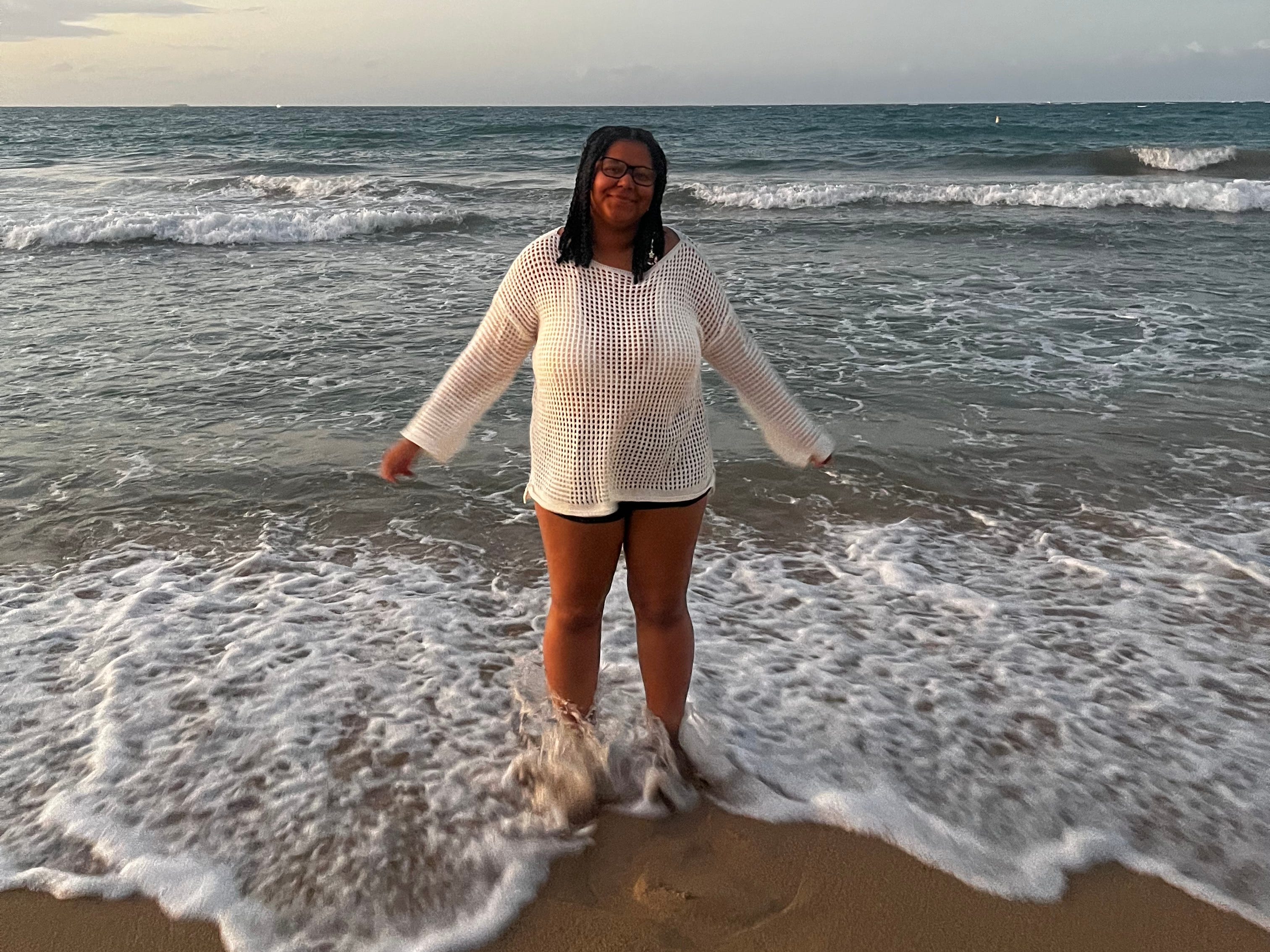 The author posing at Condado Beach at sunset. She's wearing a white long-sleeved mesh top and black shorts.