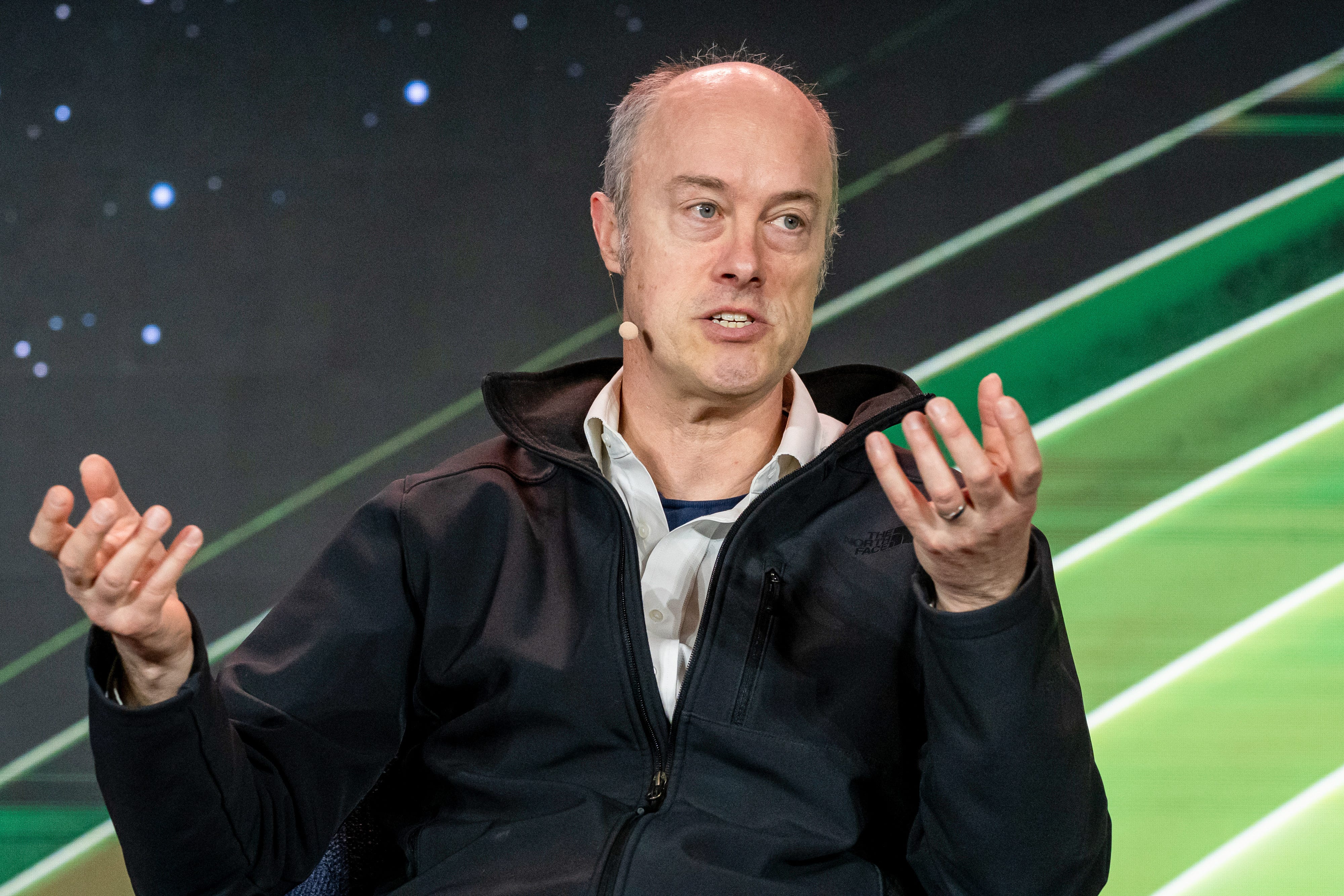 A white, bald man (Ian Buck) gestures with his hands while wearing a microphone below his ear, a light, collared shirt and a black zip-up jacket.. He sits on a stage in front of a striped green and black background.