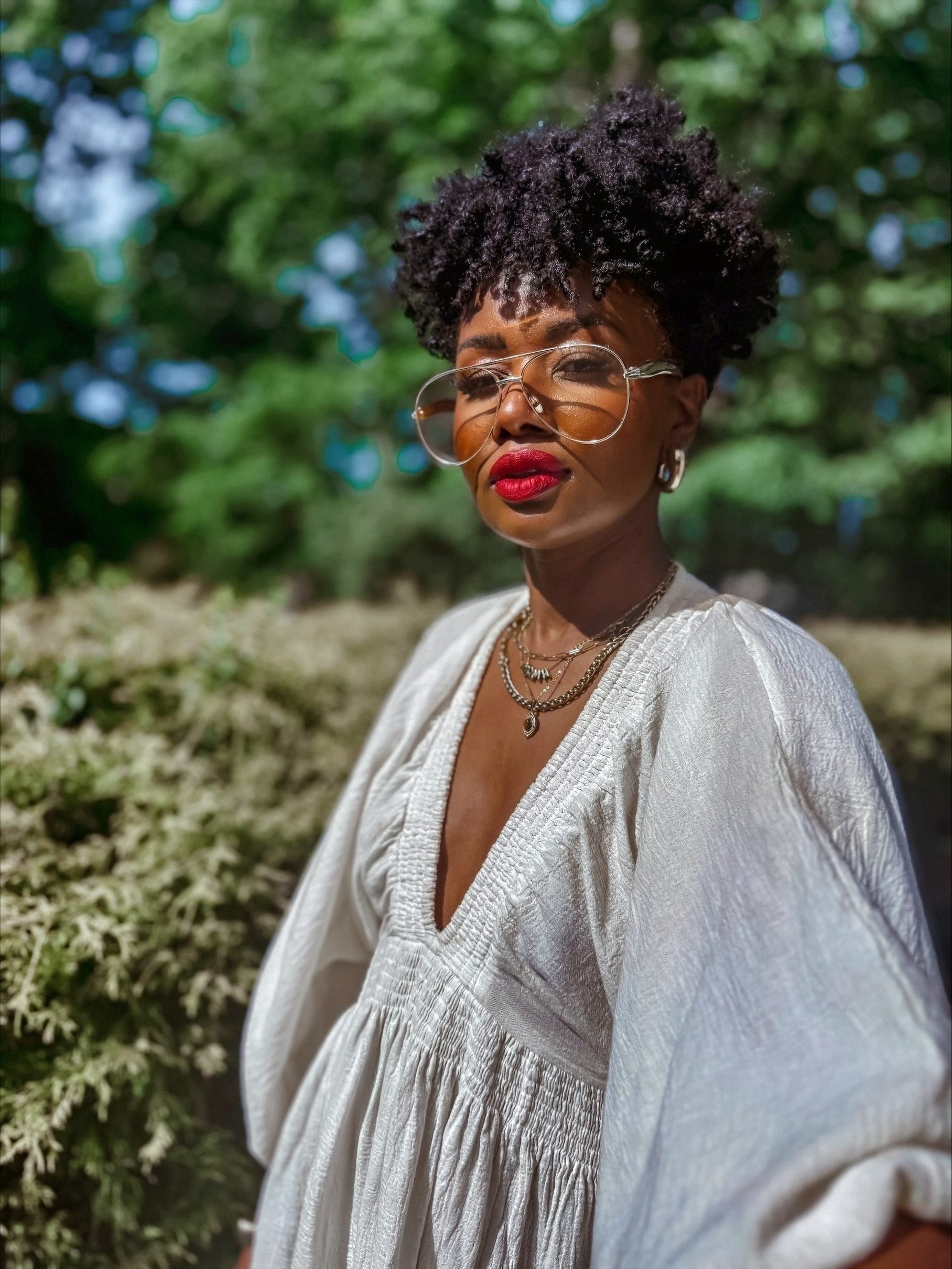 woman in red lipstick, jewelry, and glasses posing in front of a blurred background of trees.