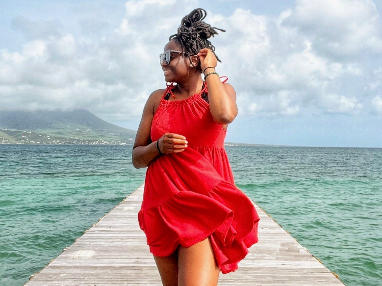 Mariette, wearing a red dress and sunglasses, walks along a pier on a cloudy day. The ocean is a beautiful turquoise color.