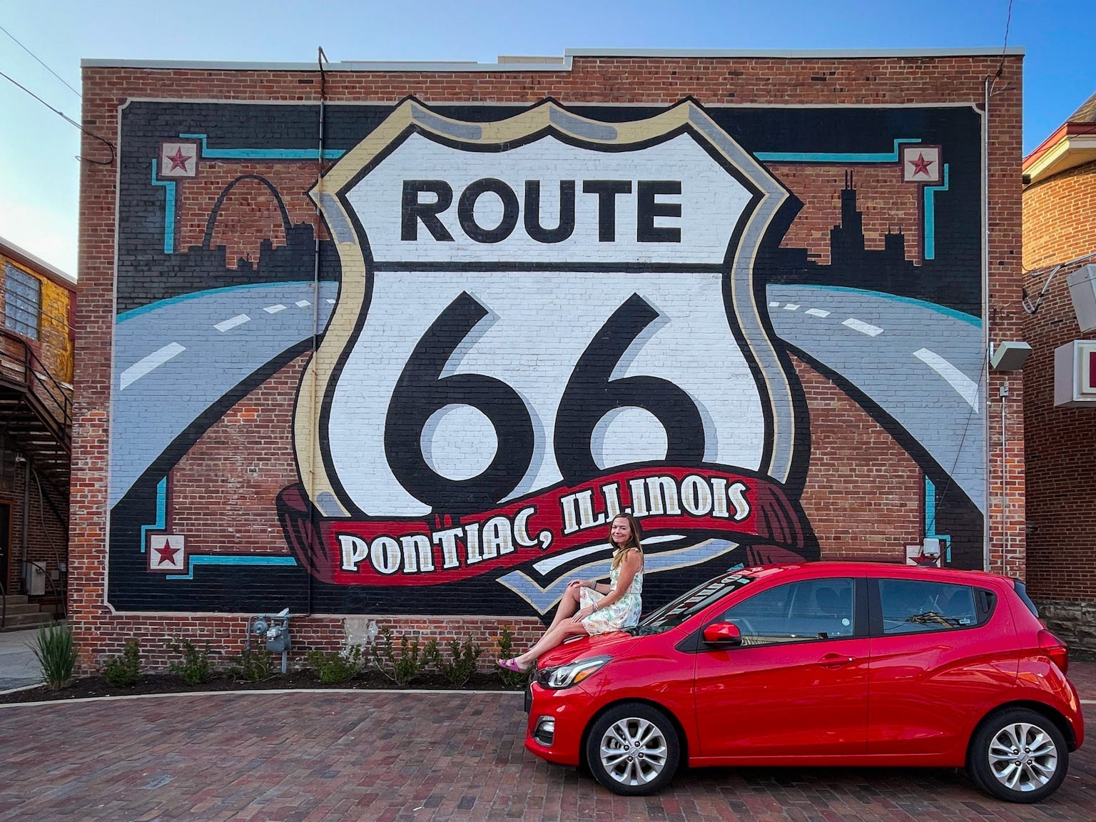 Emily sits on the hood of a red car in front of a mural that reads "Route 66 Pontiac, Illinois."